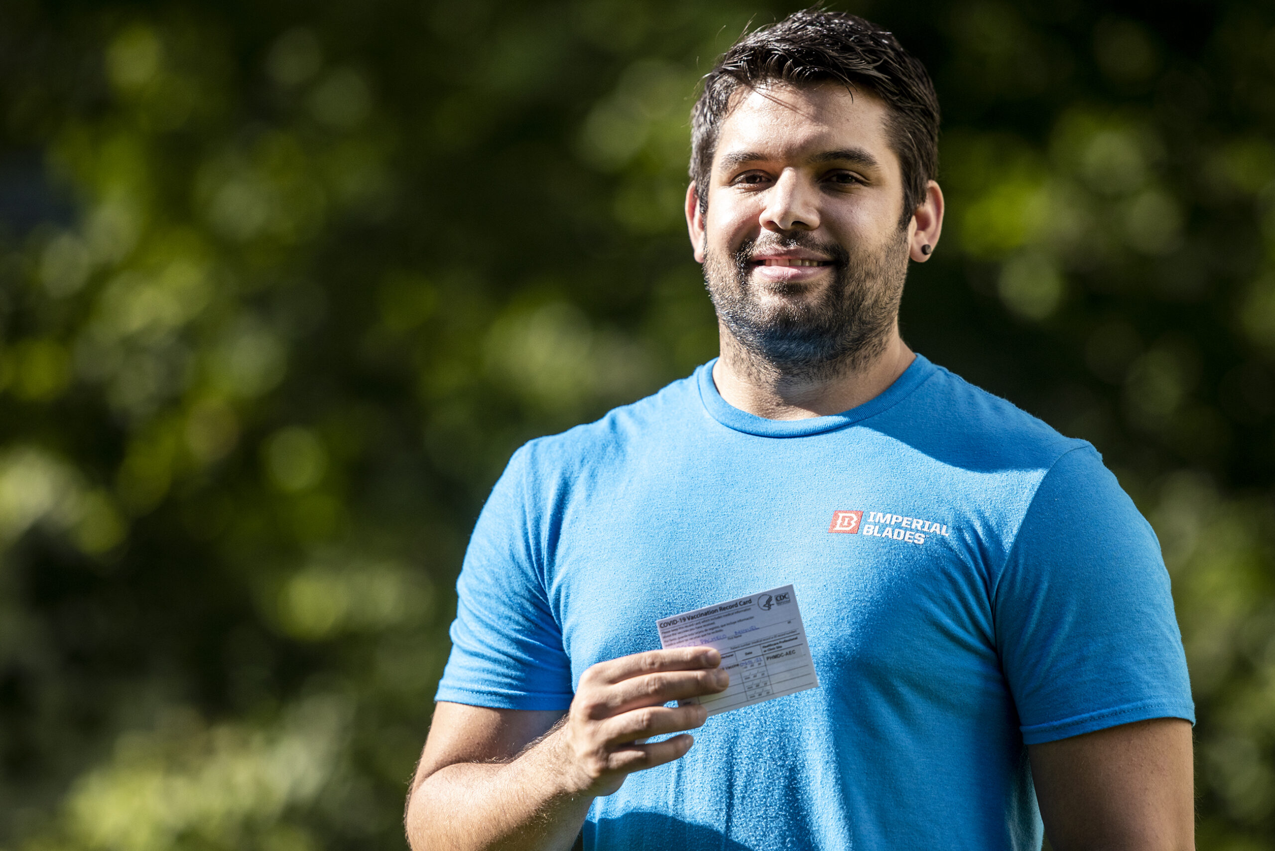 A man in a blue t-shirt holds a white card containing vaccine information.