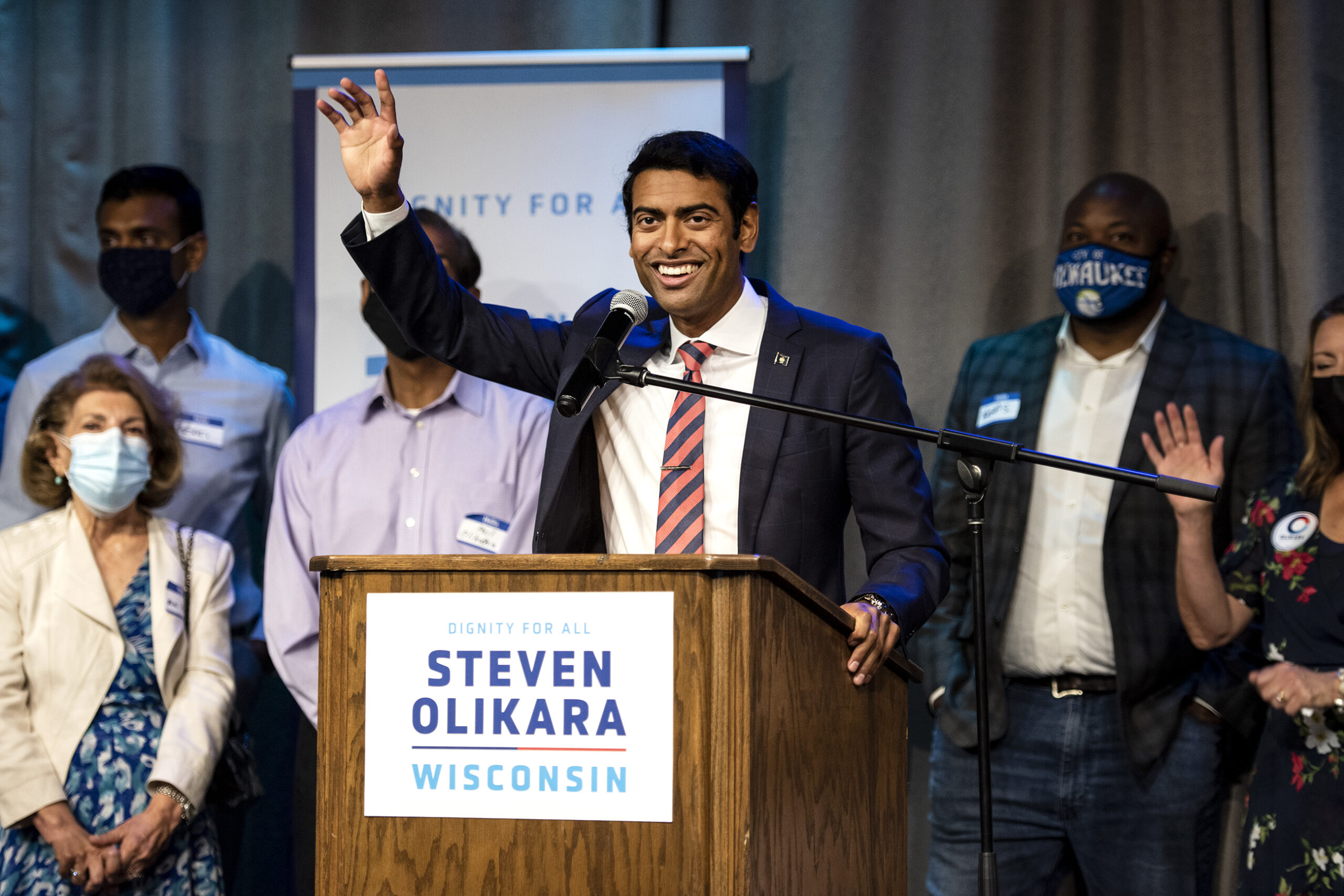 Steven Olikara smiles and waves from the podium. Supporters stand behind him.