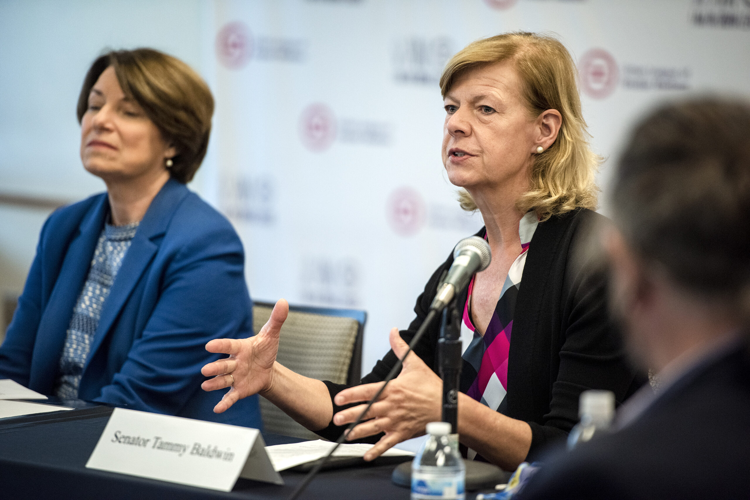 Sen. Baldwin sits at a table with microphones as she speaks to reporters.