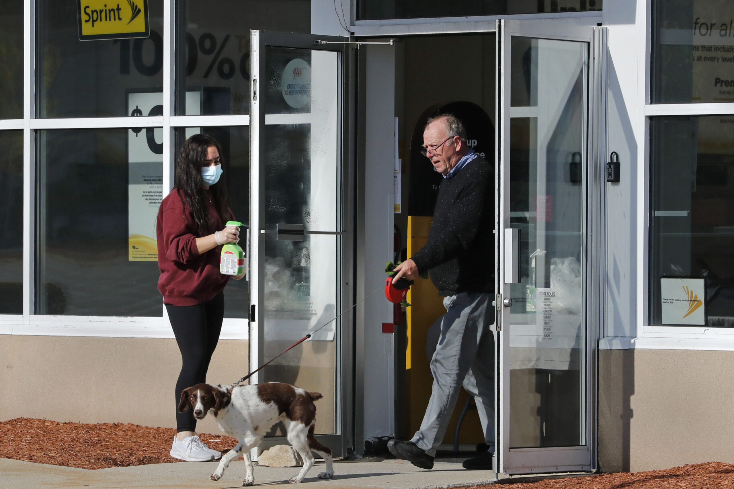 A masked worker a Sprint mobile phone store sanitizes a door as an unmasked customer walks a dog out of the store