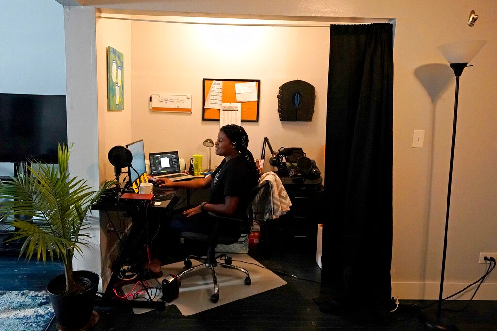 A teacher sits at her desk with a mic, laptop and several screens in her own home.