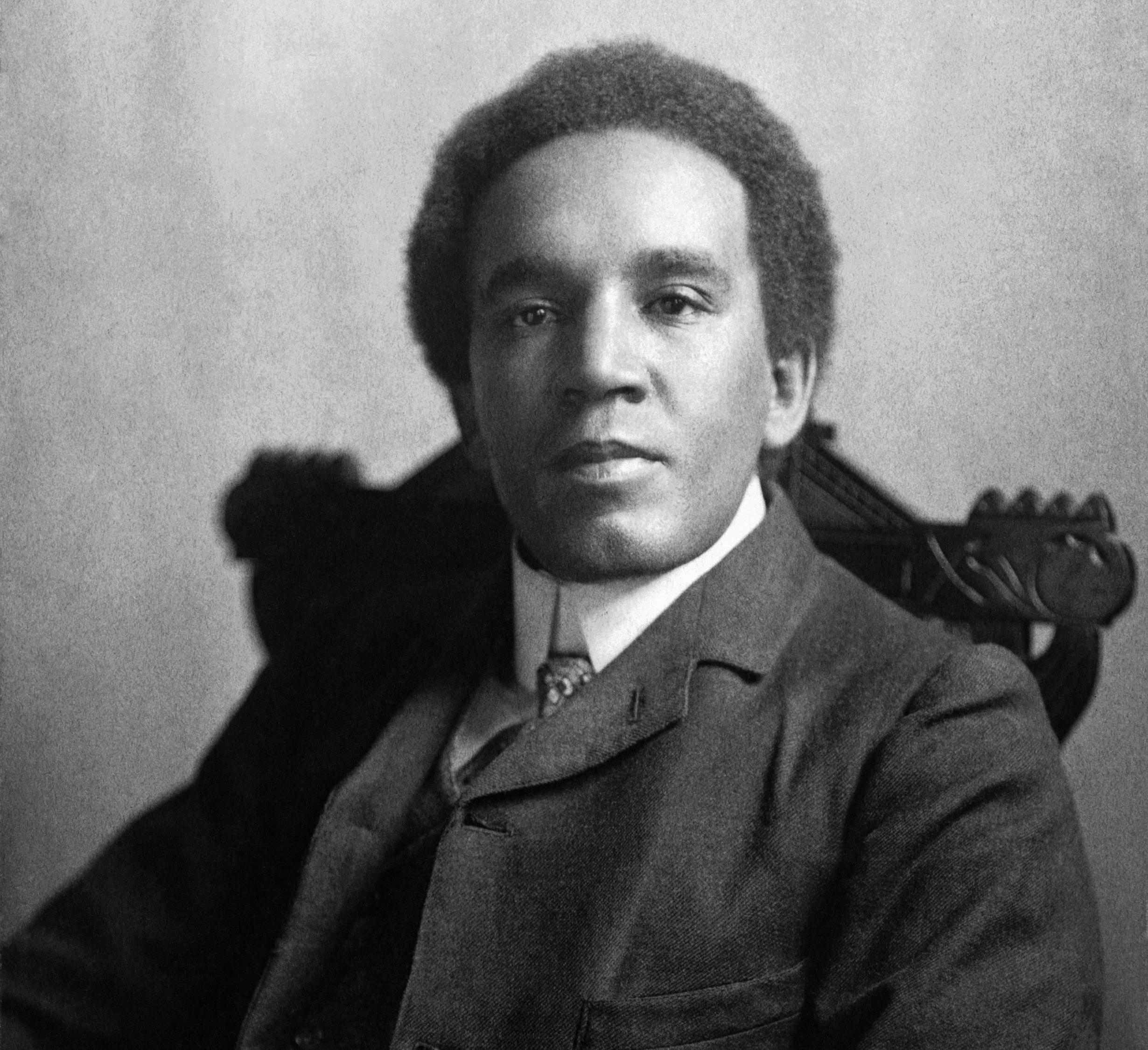 An old black and white photo of a Black man sitting in a chair, looking directly at the camera
