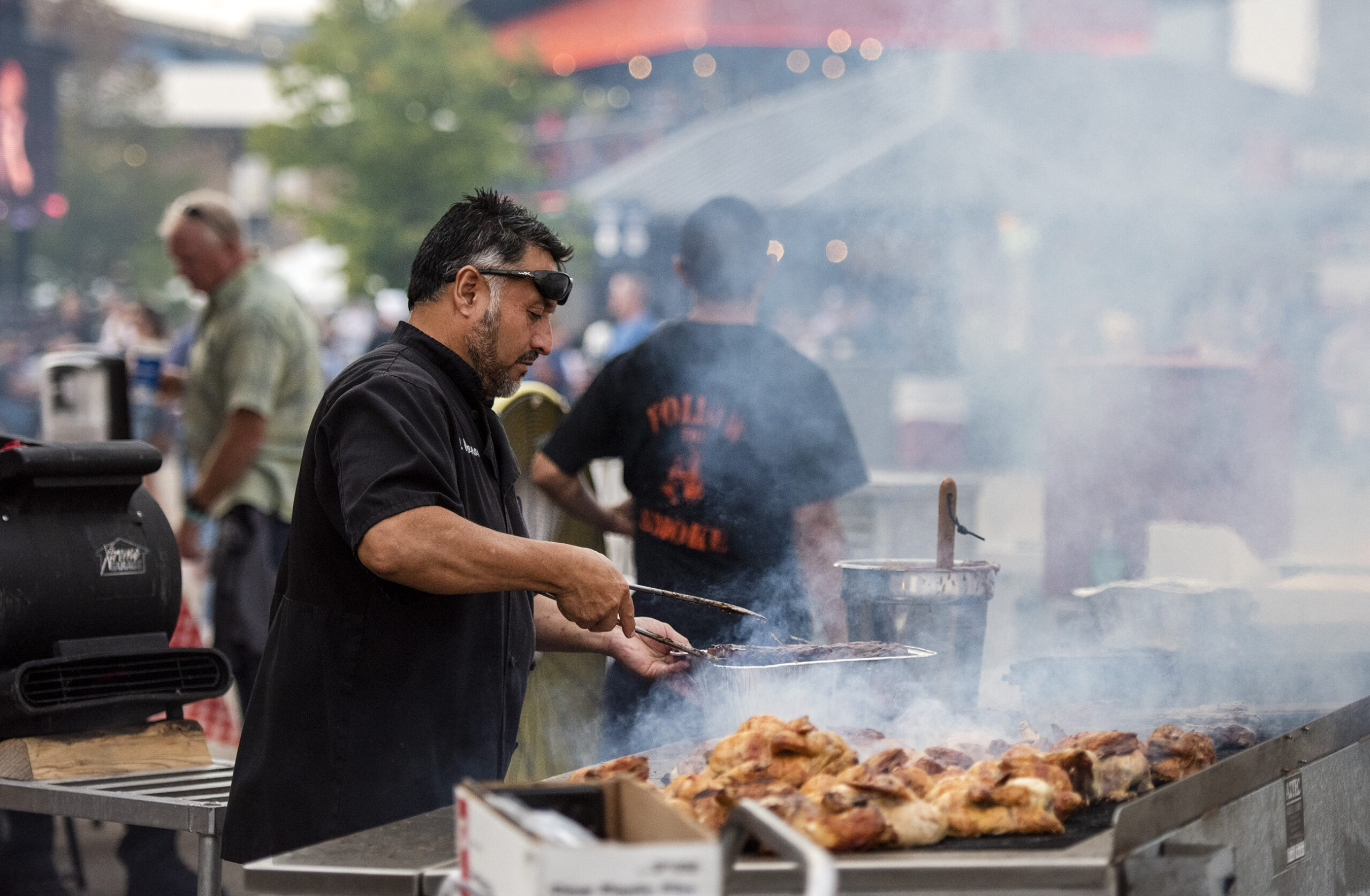 Smoke fills the air as a man prepares chicken on a large, flat grill.