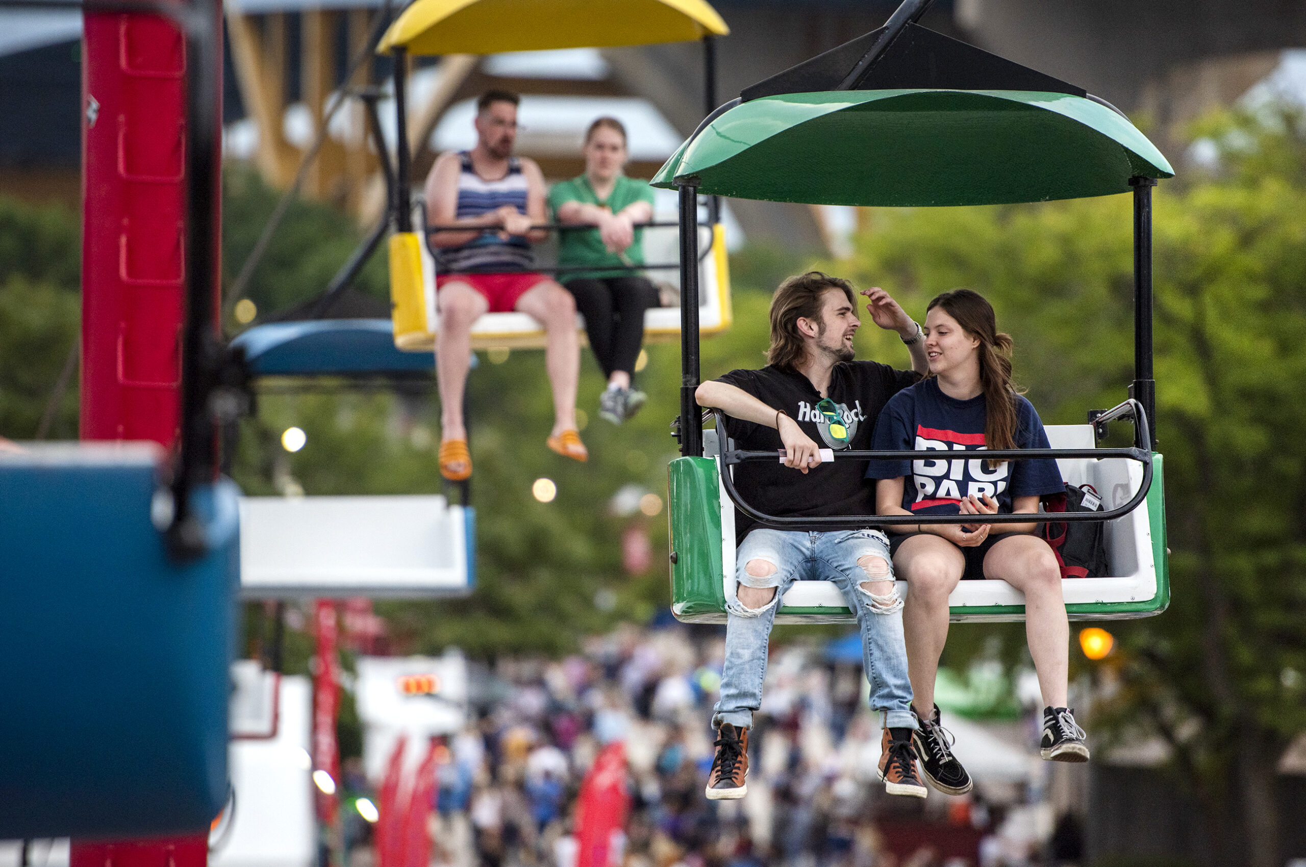 A man and a woman sit in a green SkyGlider seat as they coast above the crowd below.