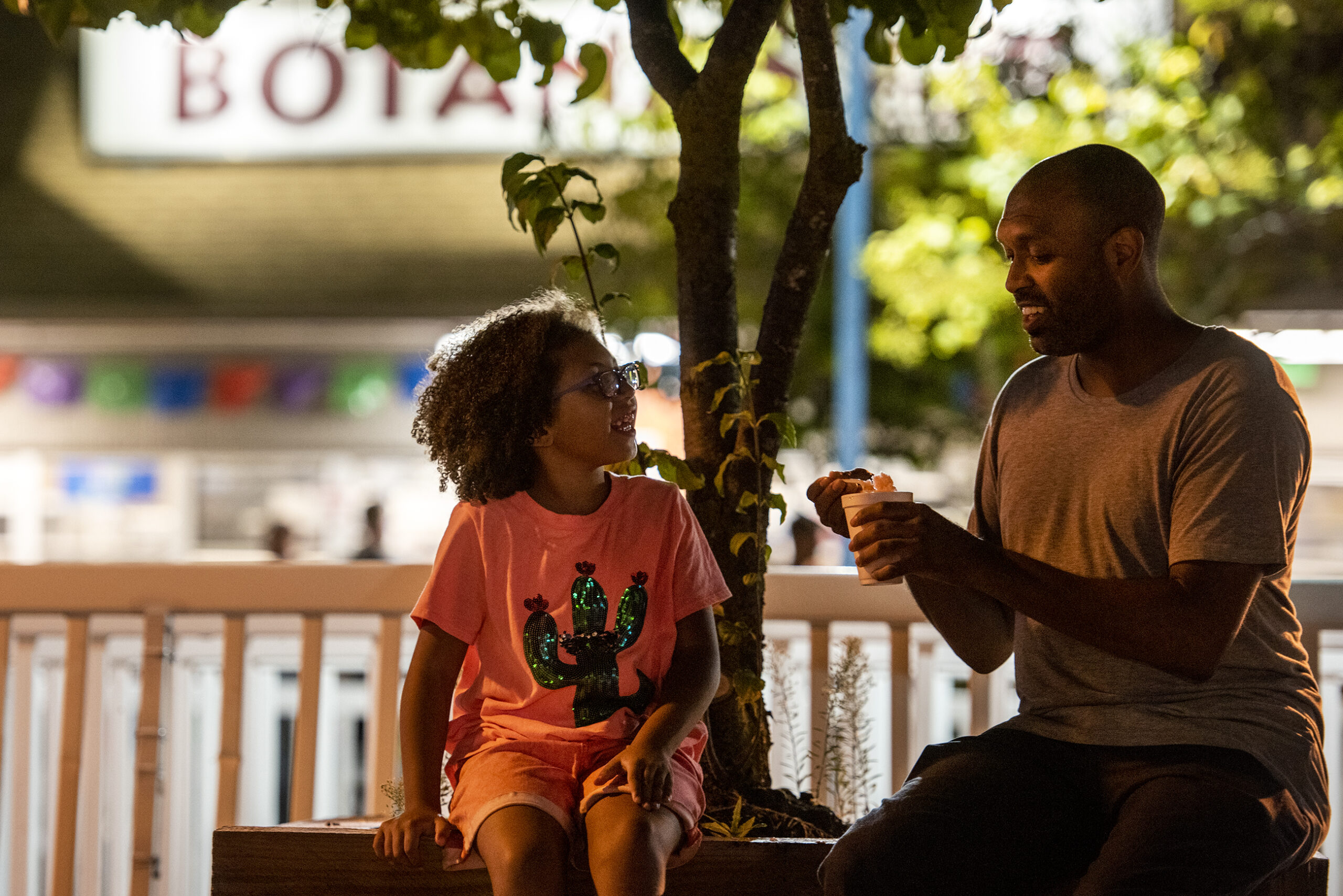 A father and daughter sit on a bench as they eat shaved ice.