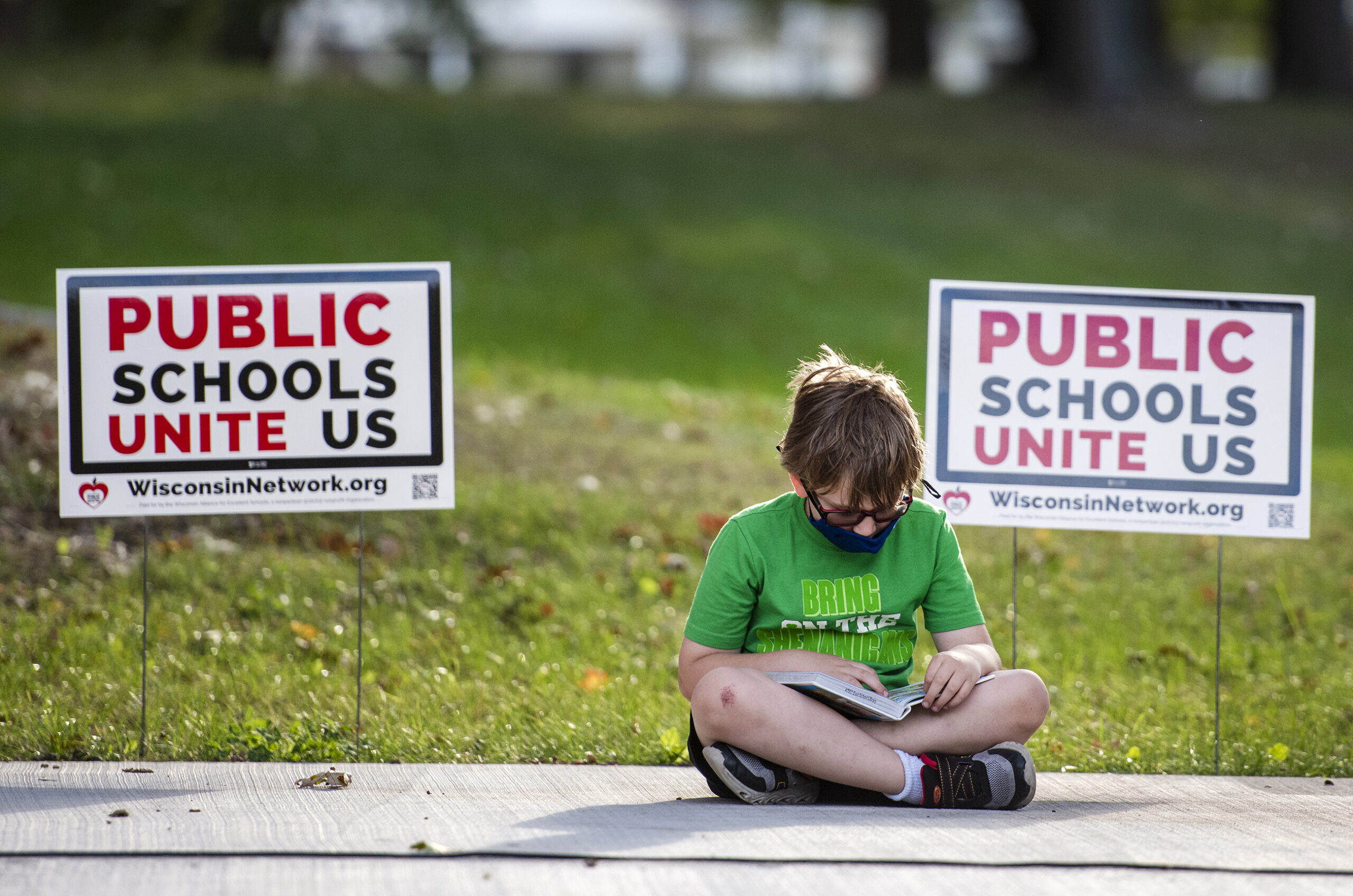 A child in a green t-shirt sits on the ground outside with his head in a book.