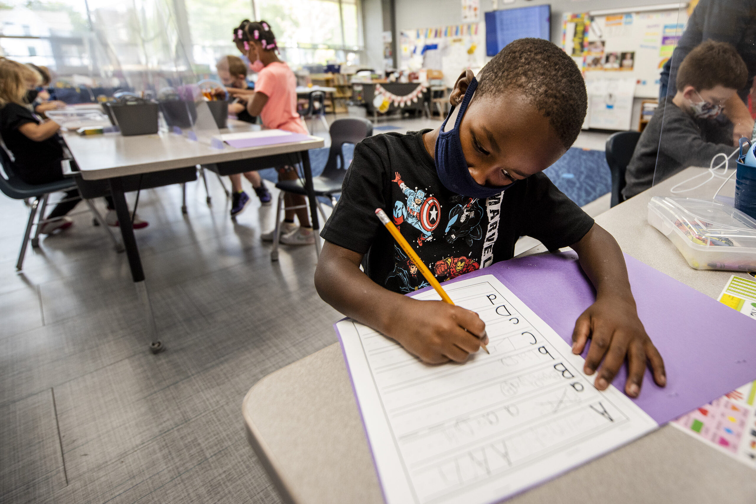 A student writes with a pencil as he wears a face mask at his desk.