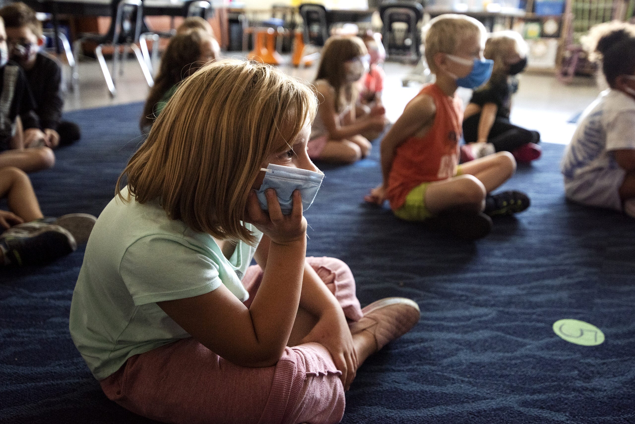 A girl in a face mask rests her head in her hand as she listens to a story being read.