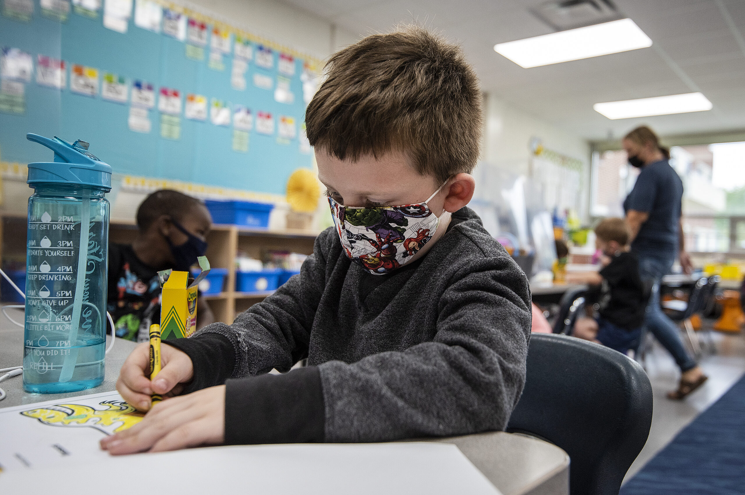 A boy in a face mask uses a crayon to color a picture.