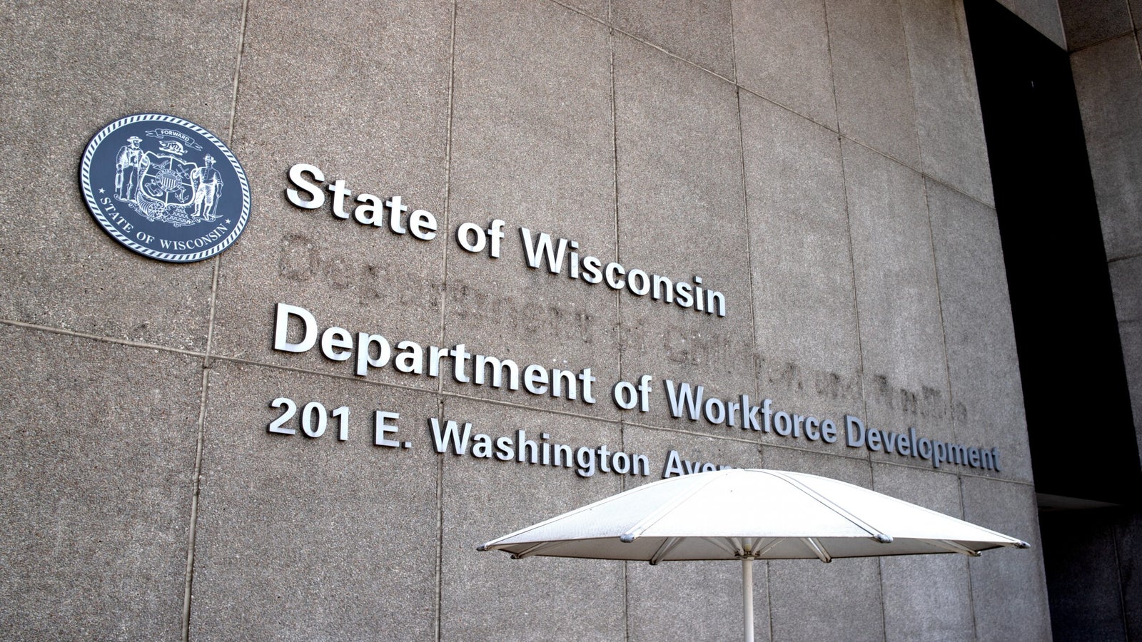 Building exterior with State of Wisconsin Department of Workforce Development, 201 E. Washington Avenue sign and state seal, partially shaded by a white umbrella.