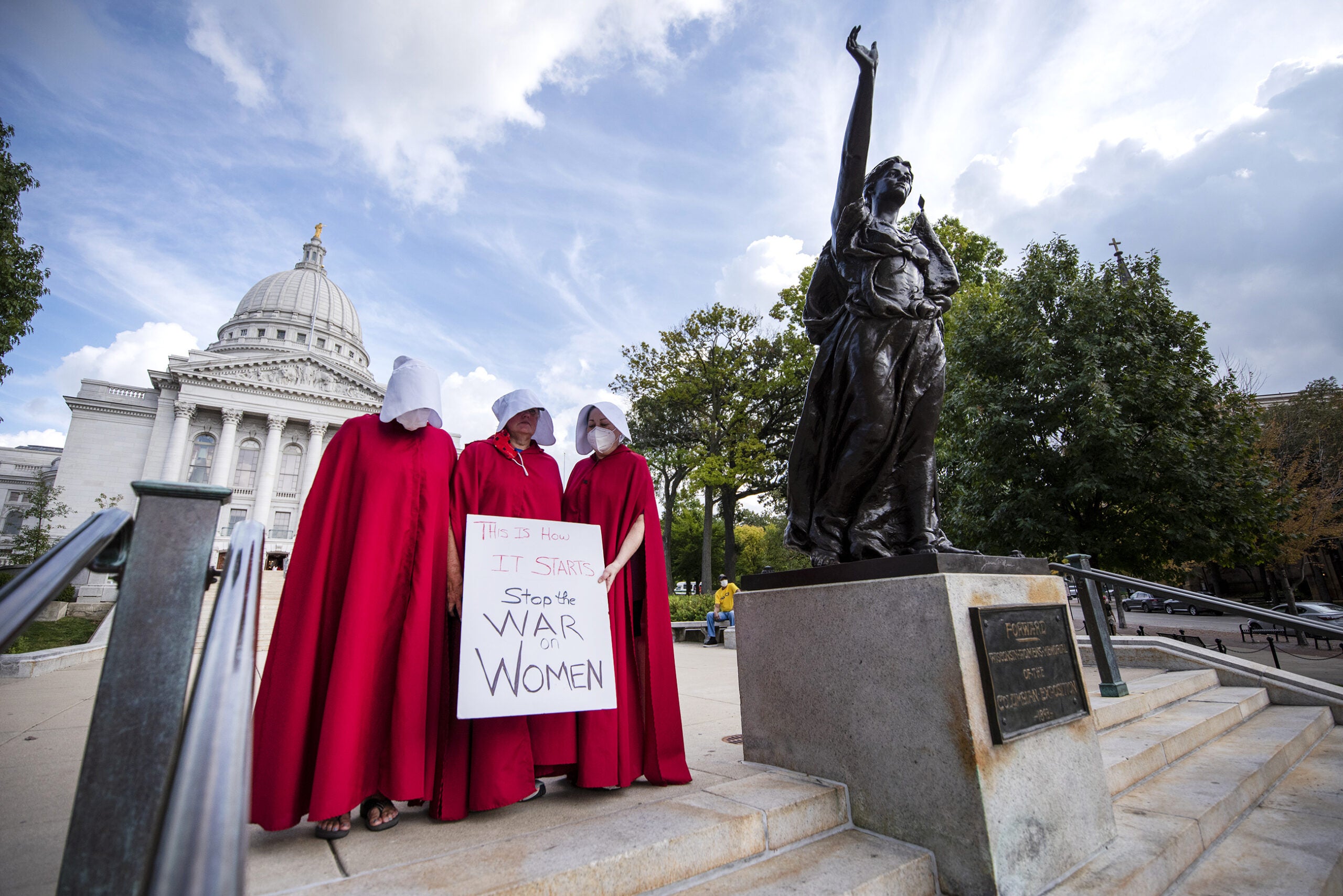 Three people in "Handmaid's Tale" costumes consisting of red robes and white bonnets stand near the state capitol.