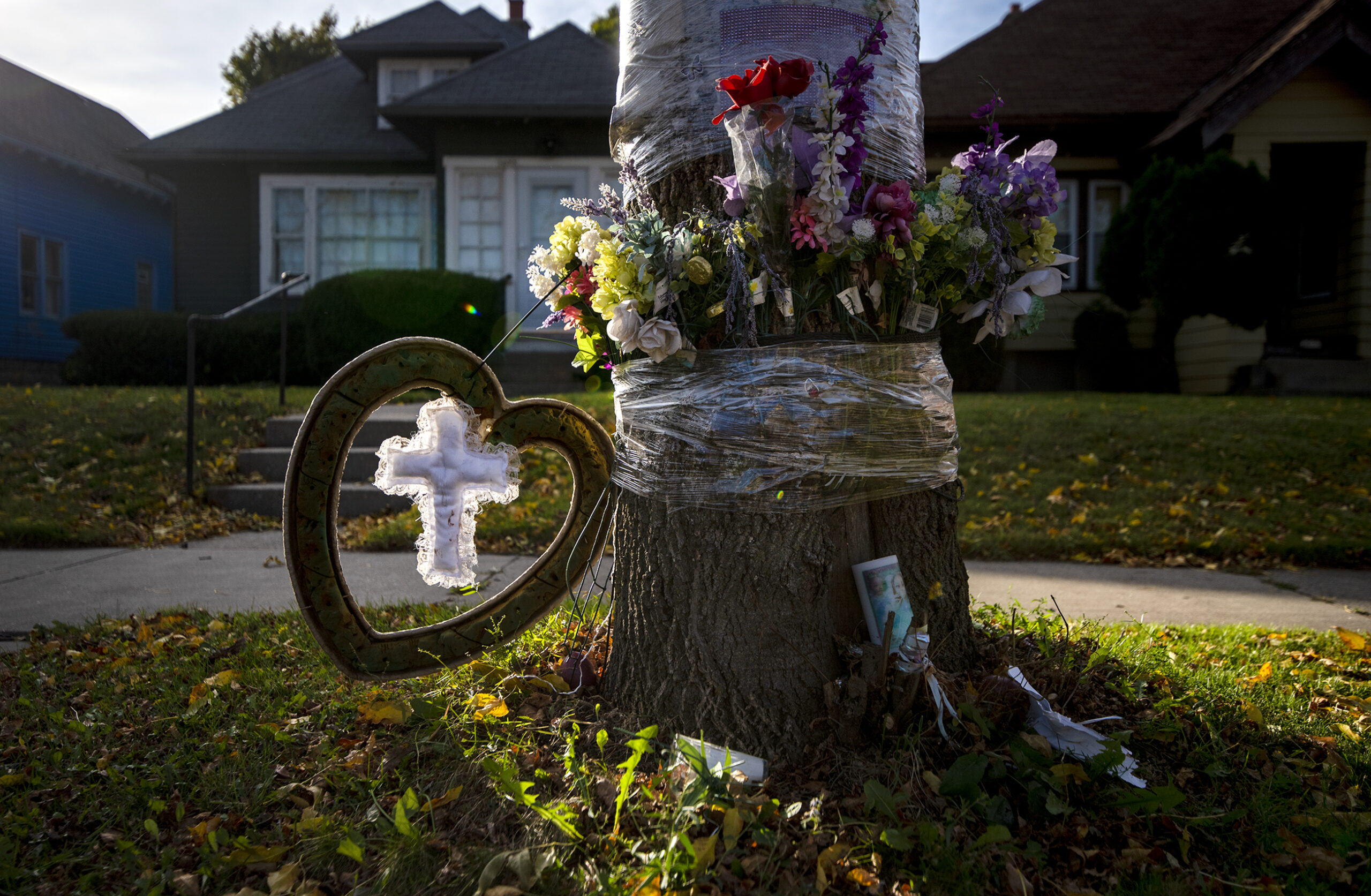 A white cross hands inside of a heart shaped wreath along with flowers and other memorial items on a tree.