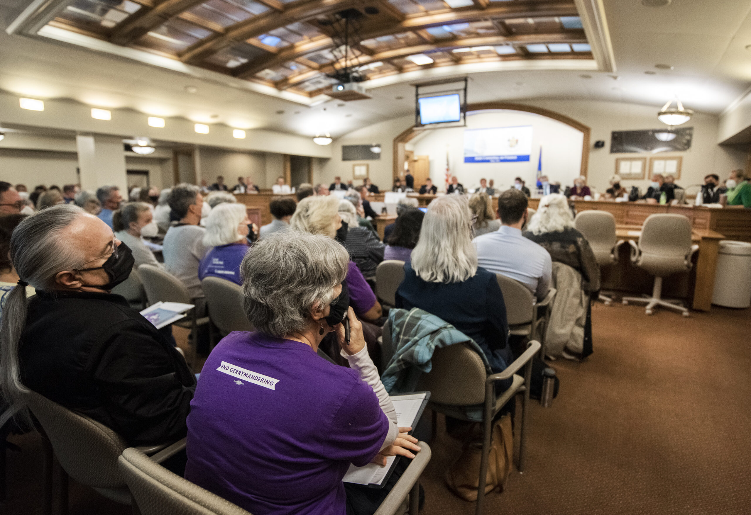 An attendee in the back of the room leans against the arm rest as she listens to state legislators speaking at the front of the room.