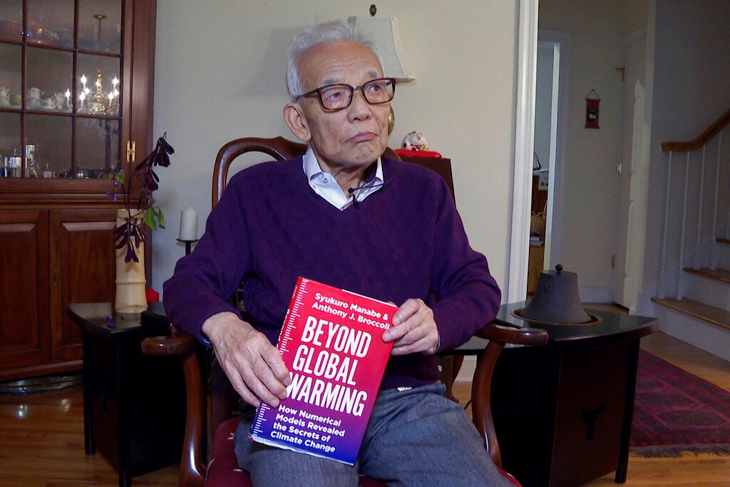 A man wearing glasses and purple sweater sits in chair holding book.