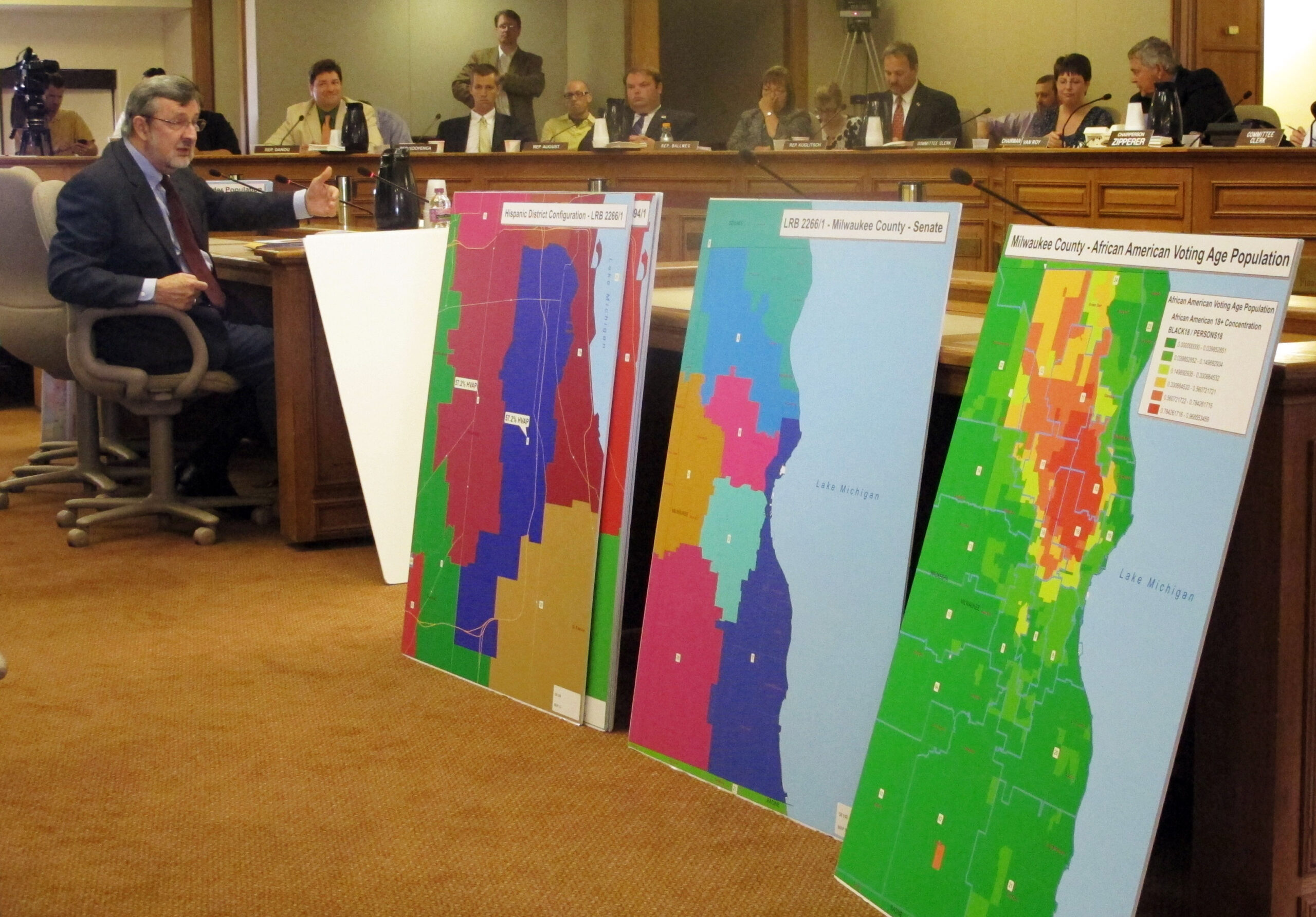 Lawmakers seated behind a hearing room desk with large poster maps in the foreground
