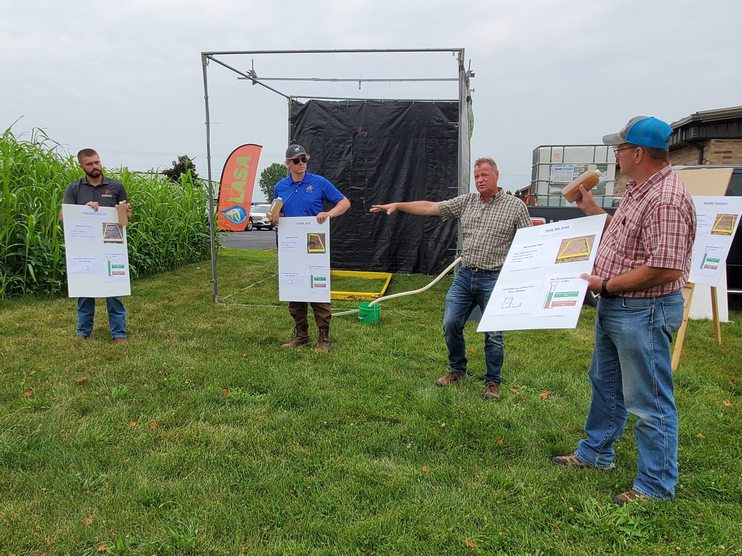 Four men stand in the grass next to a farm field with a few holding poster board.