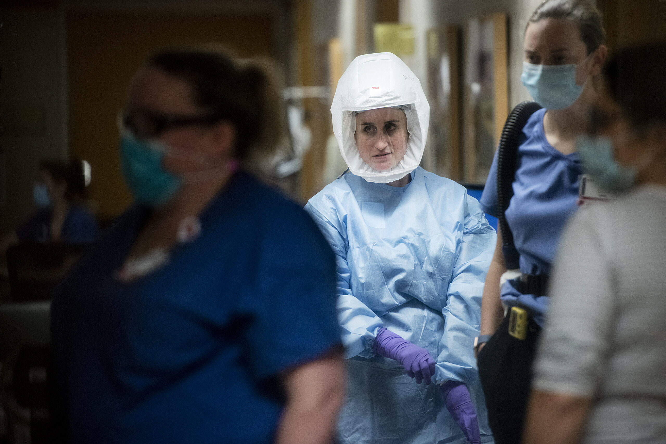 A woman in a blue gown, purple gloves, and a large white hood with a clear plastic window pulls up a glove as she walks through a hallway with other healthcare workers