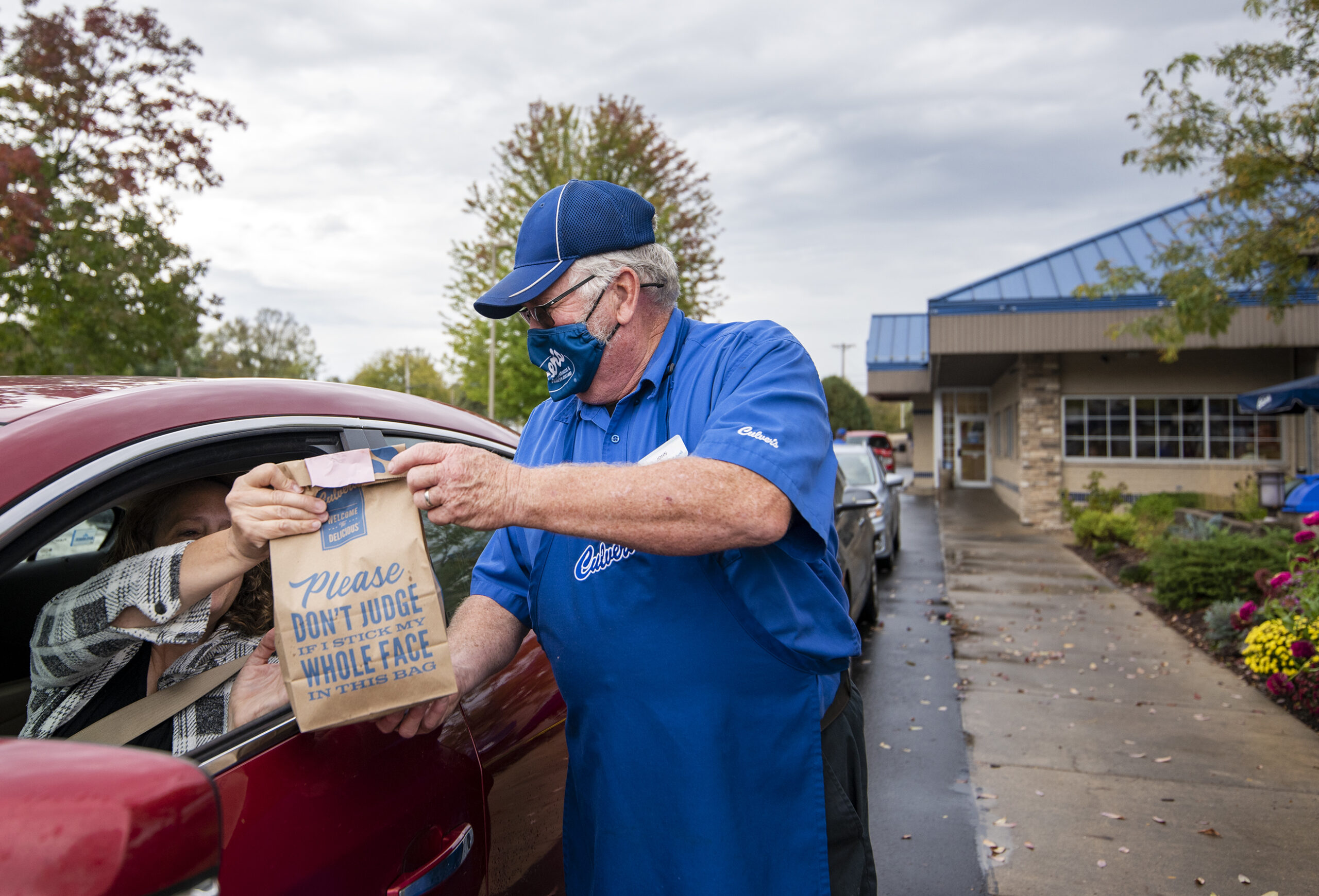 A worker in a blue uniform hands a paper bag with food to a driver in a red sedan outside of the restaurant.