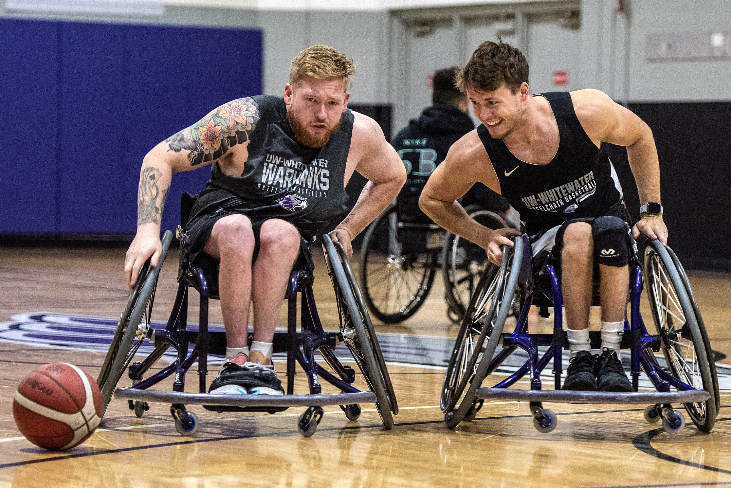 Two wheelchair basketball players in jerseys work to get to the ball.