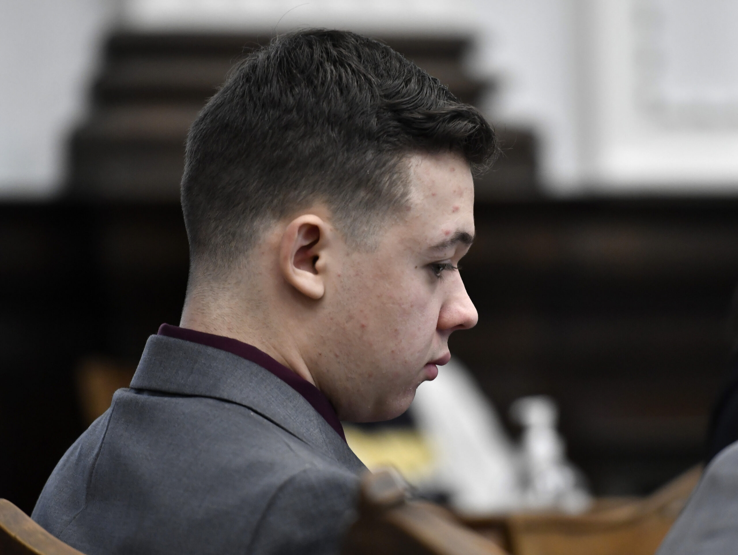 Kyle Rittenhouse in a gray suit casts his eyes at a table