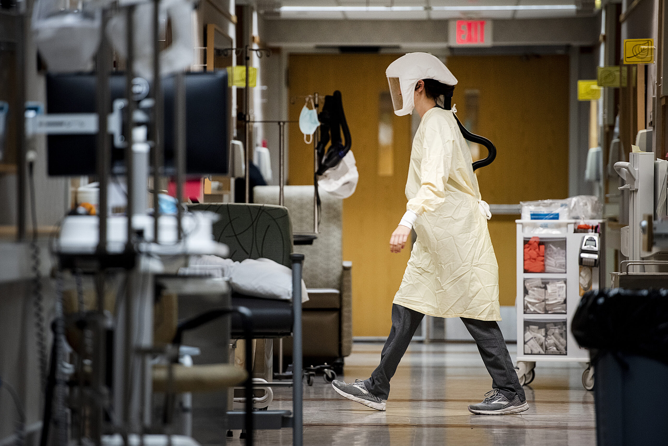 A woman in a yellow gown and protective hood walks through a hospital hallway