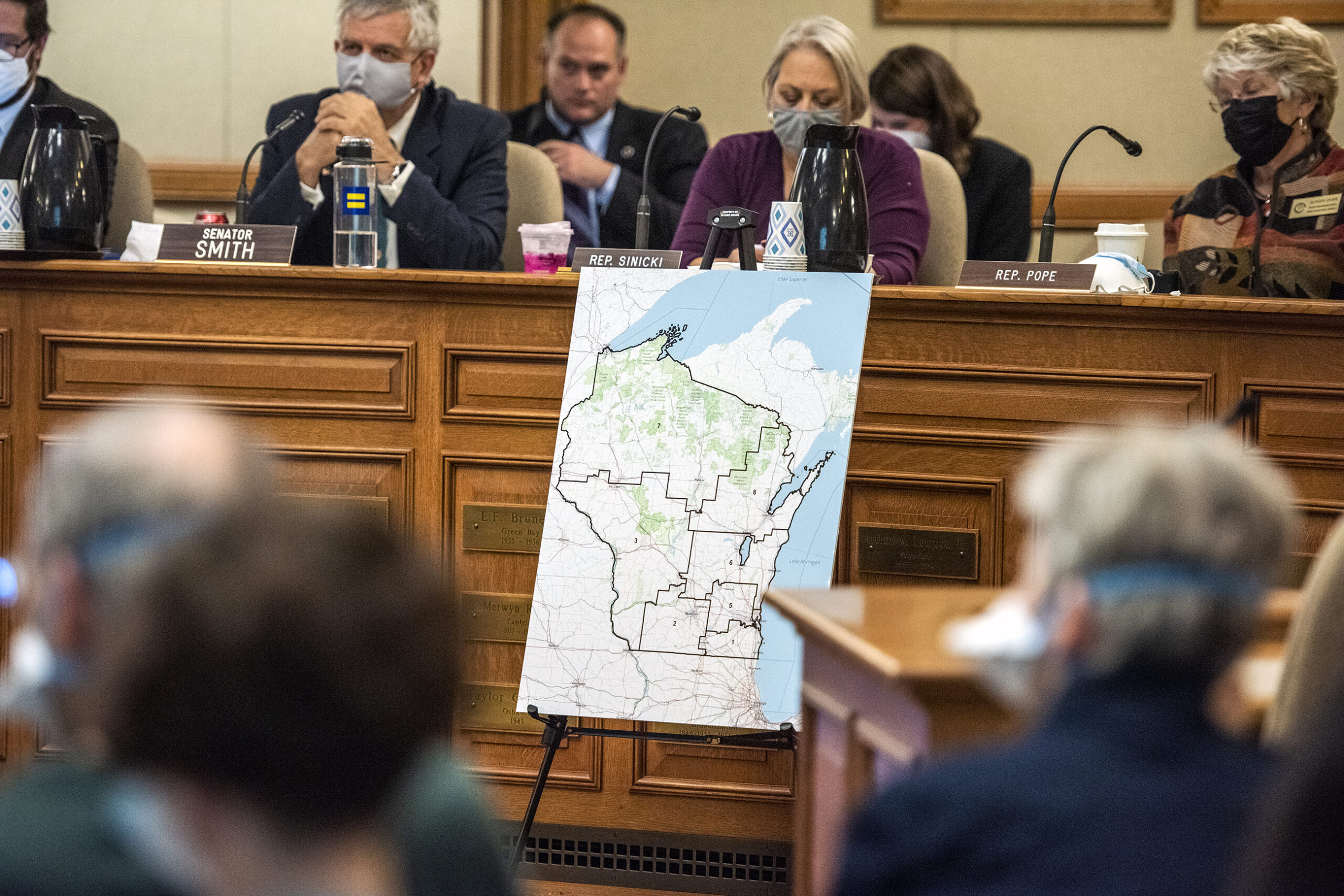 A poster showing a map of Wisconsin is displayed in front of state senators at a hearing.