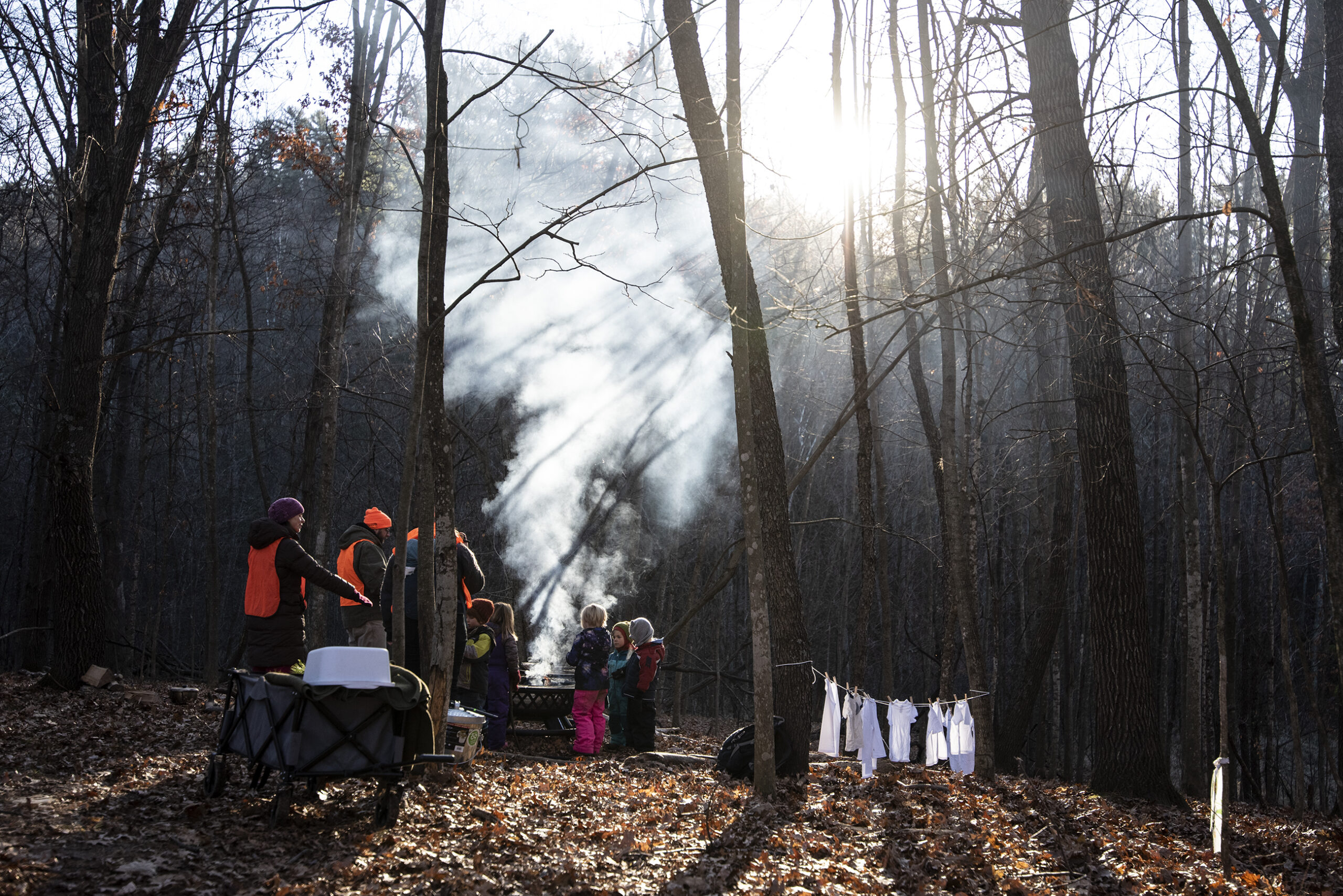 White smoke travels upwards as the sunshine illuminates it through the trees. Small children gather near the fire it is coming from.