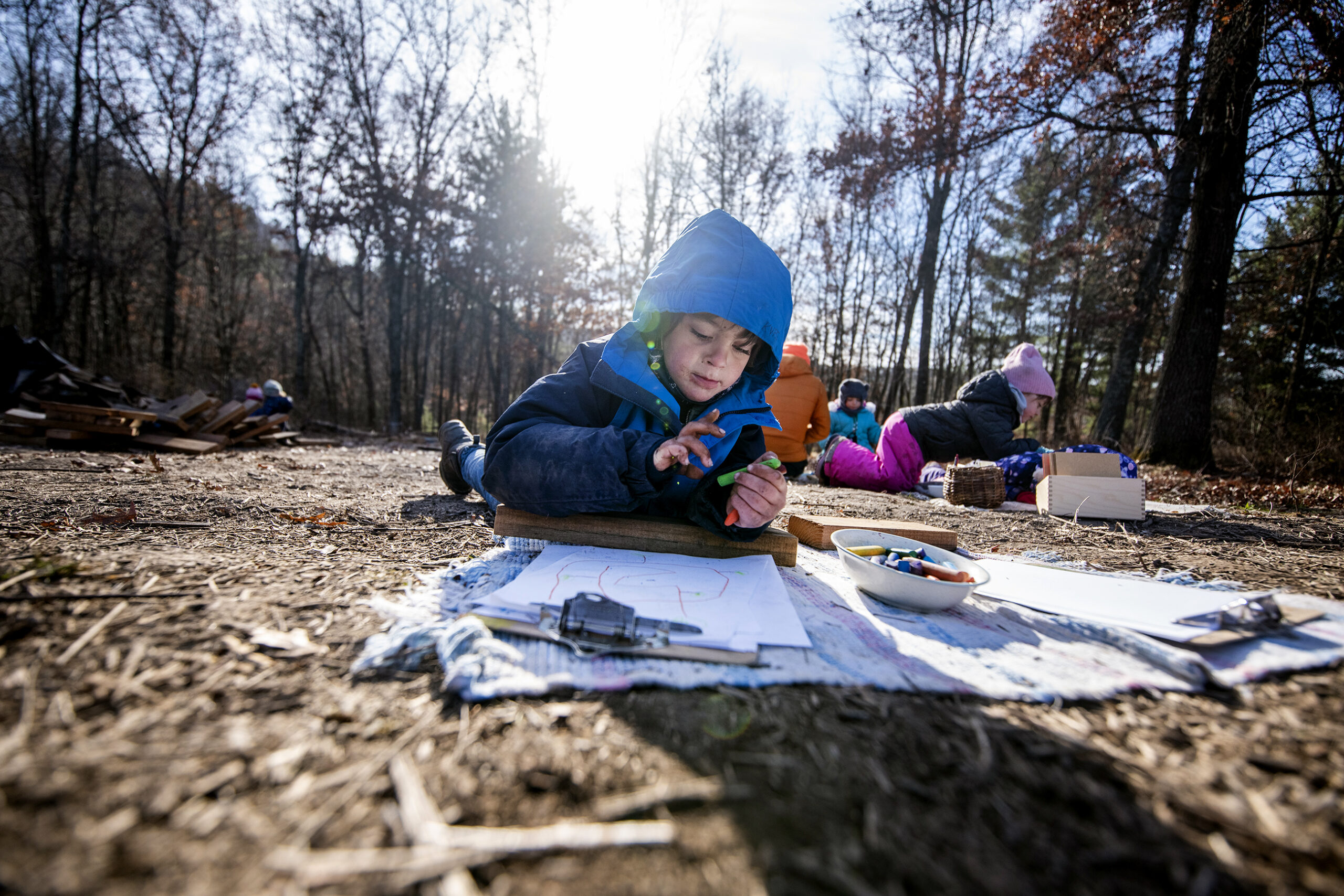 The sun shines behind a boy laying on the ground as he colors.