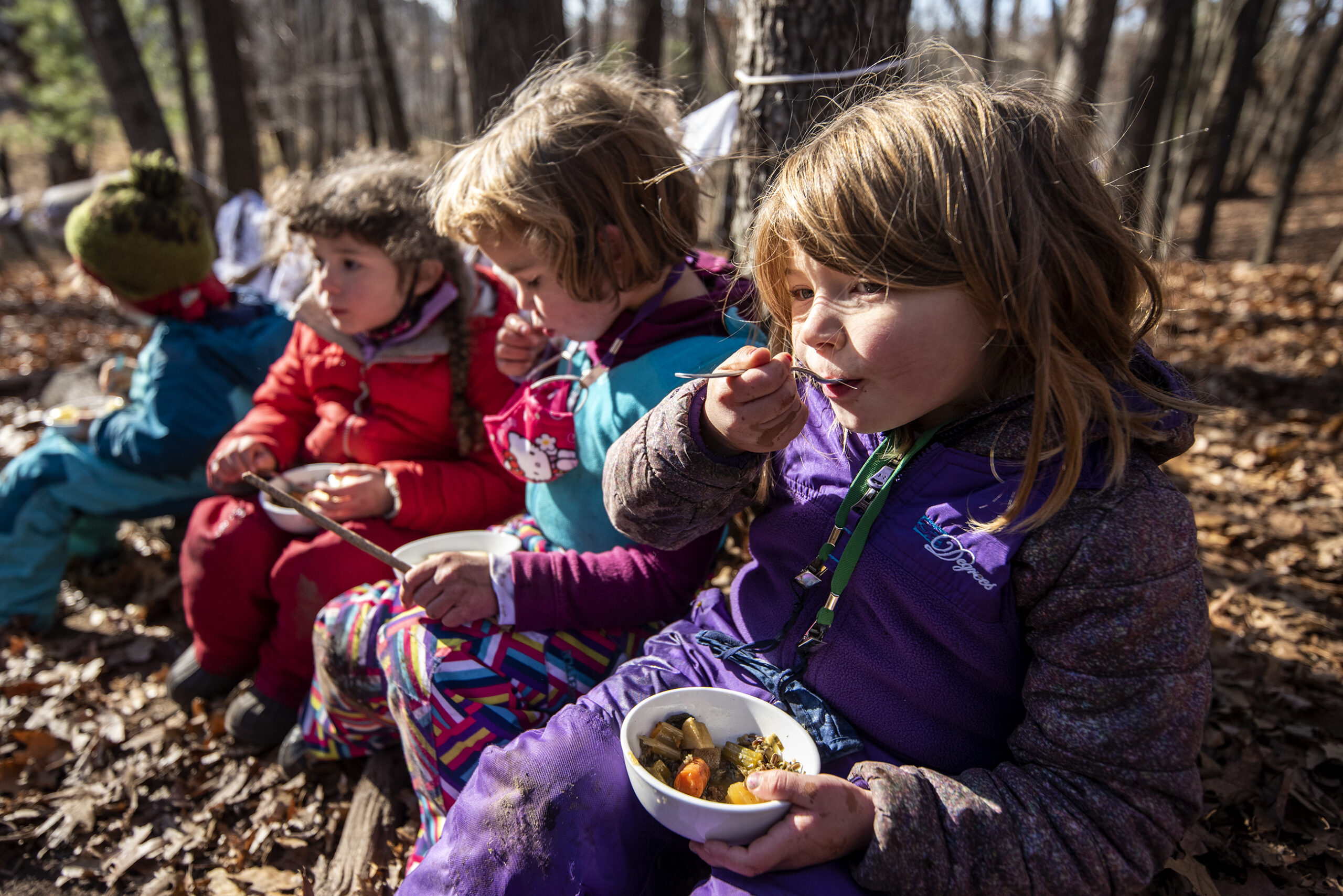 A line of students sit on a log outside as they eat soup.
