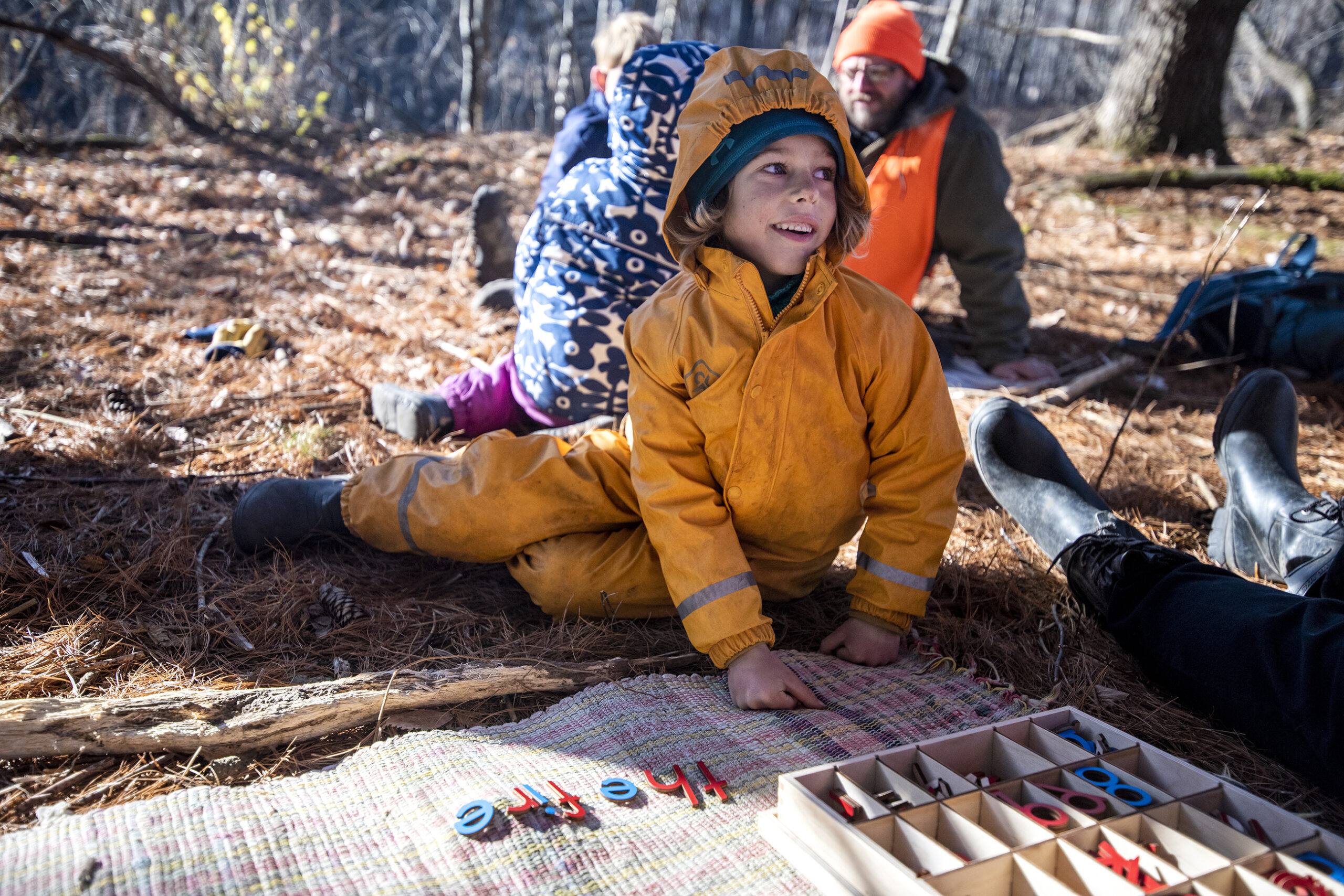 A child in a yellow winter suit sits on the ground during a spelling lesson.