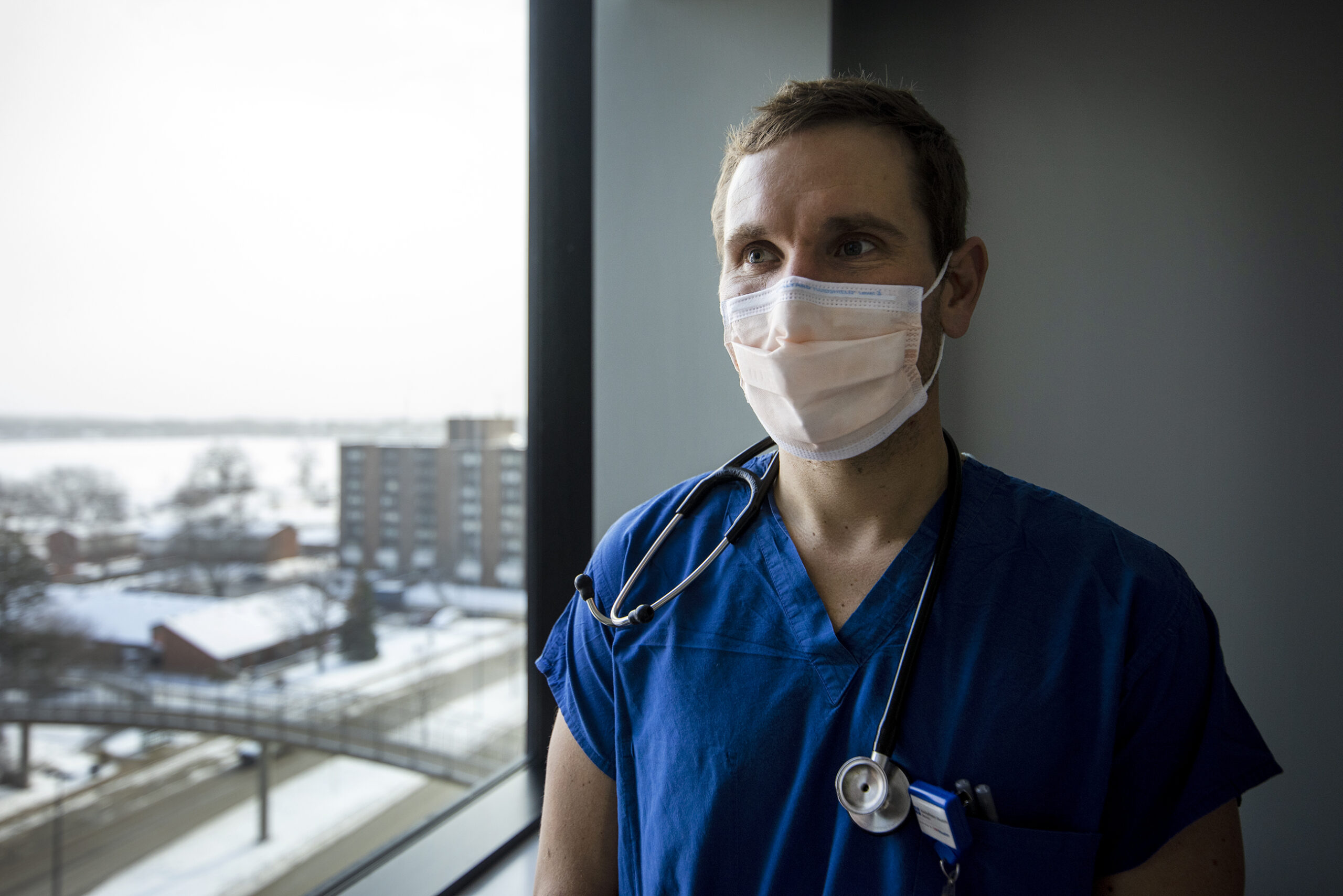 A doctor in blue scrubs and a face mask stands near a window.