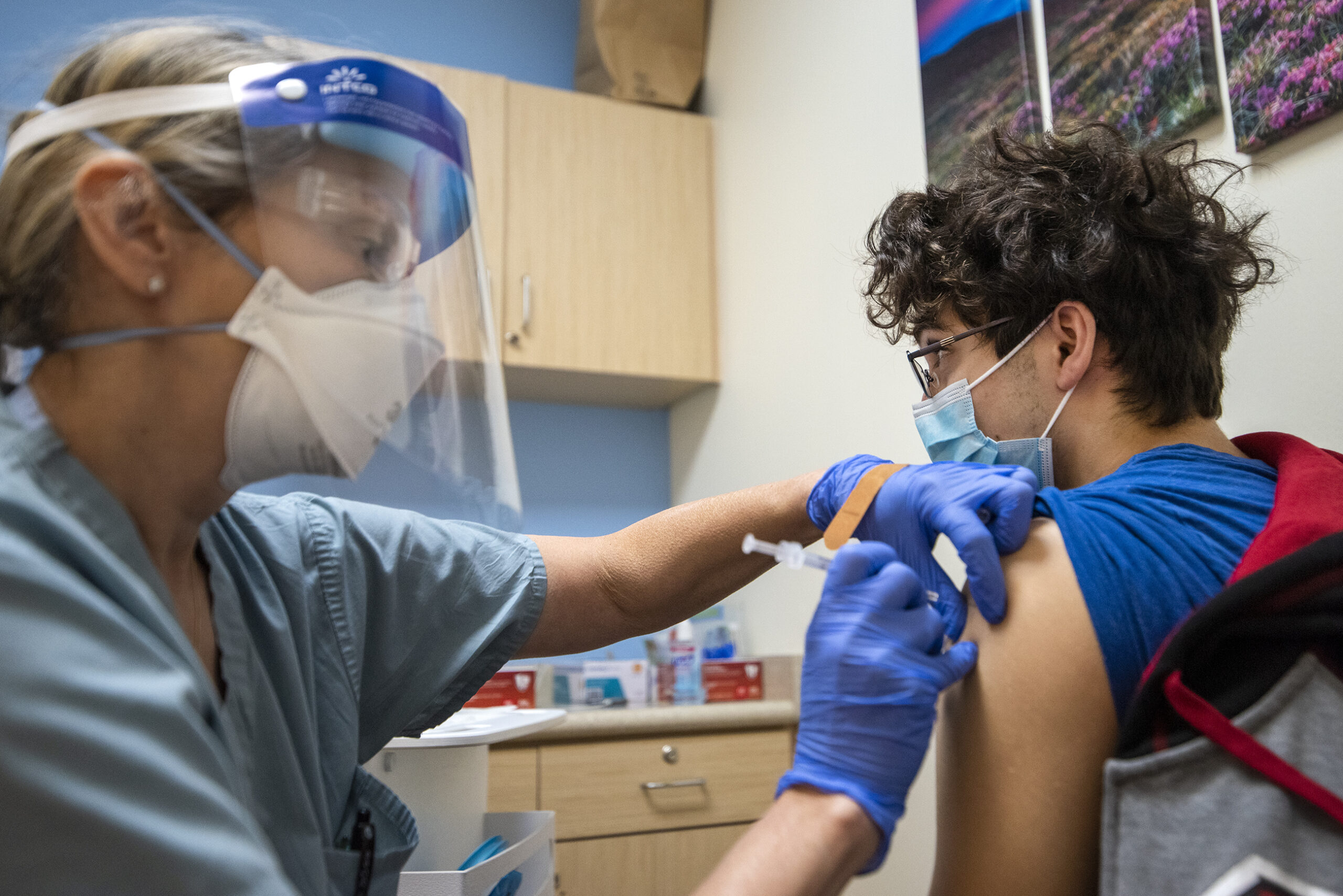 A boy in a face mask looks away as a physicians assistant injects a booster shot into his arm.