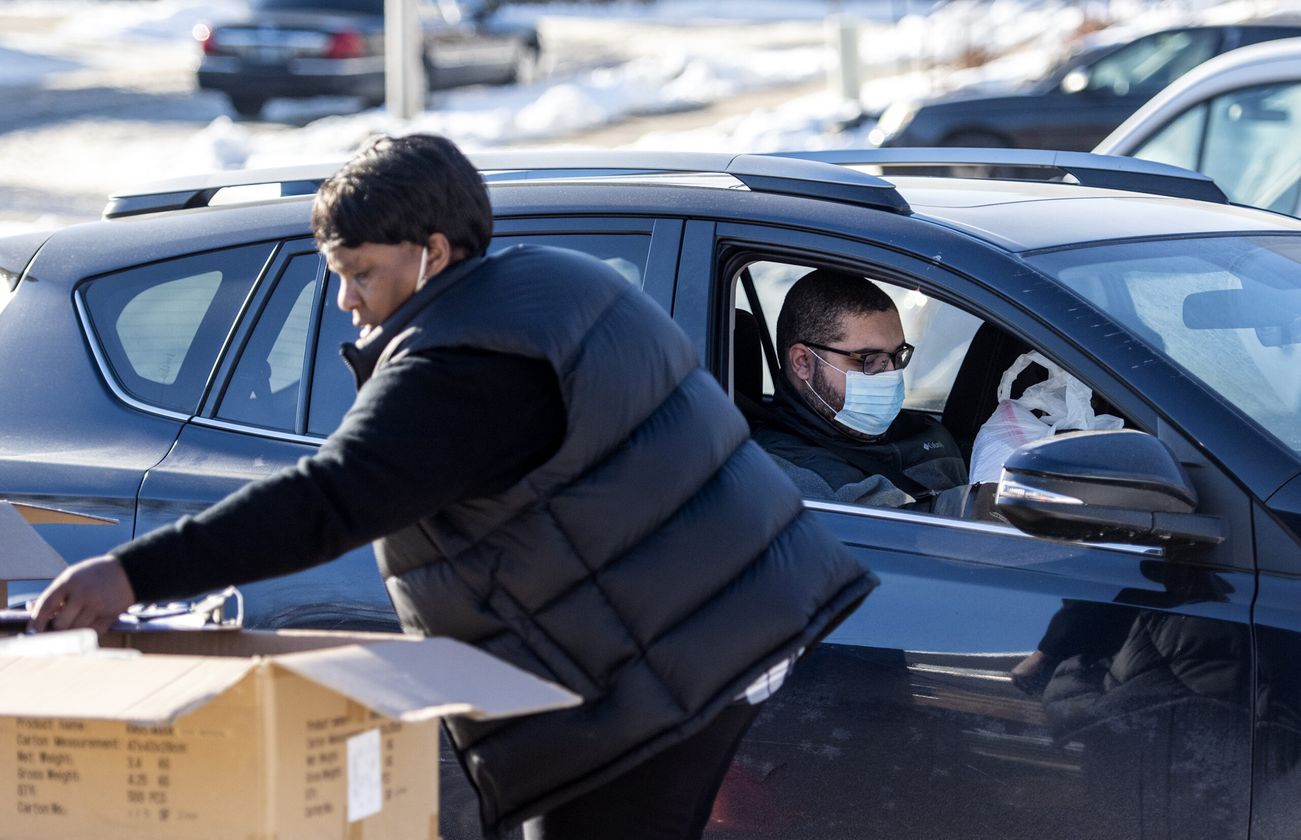 A driver wearing a face mask sits in a car as a woman delivers a bag of masks to him.