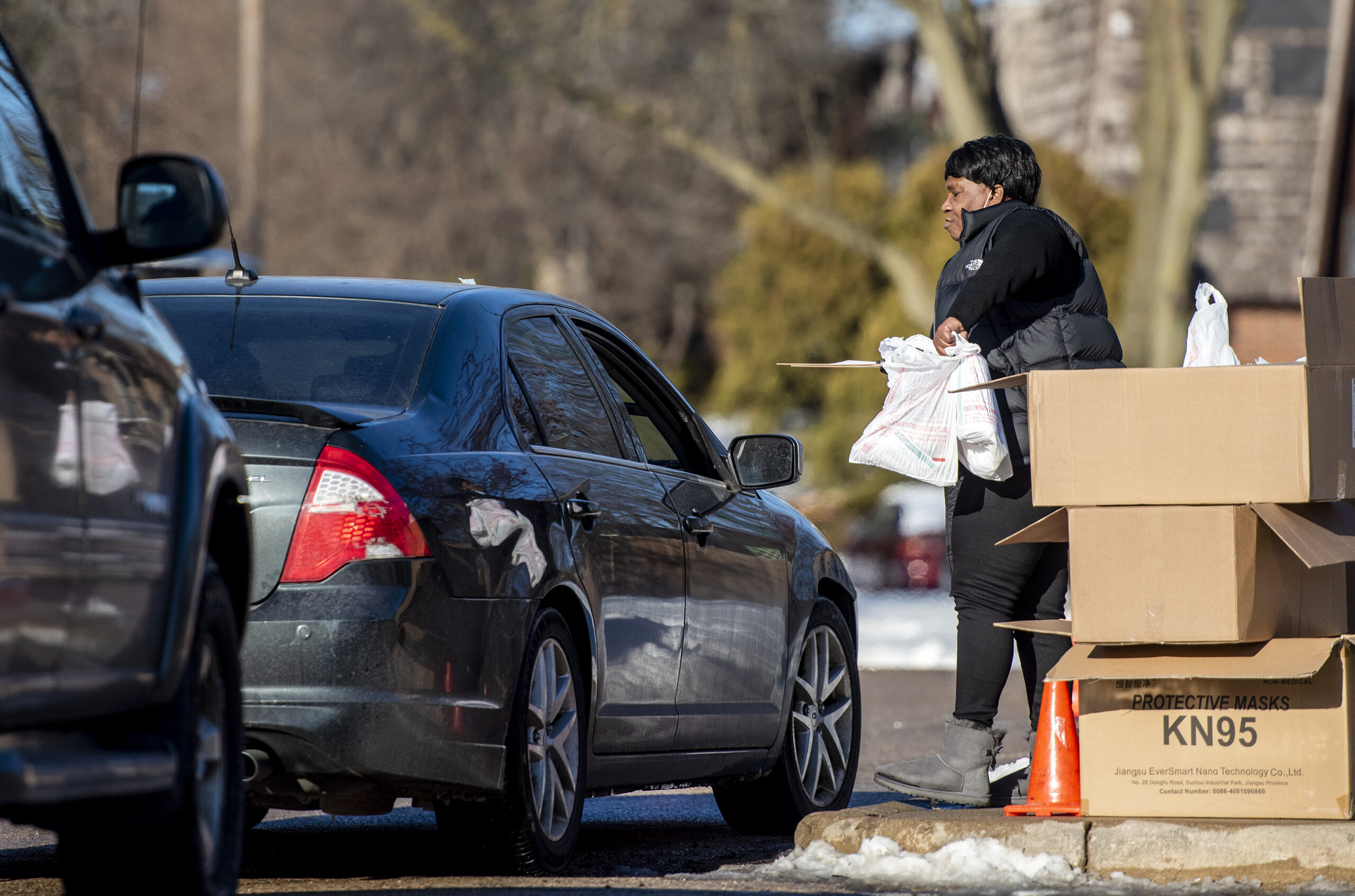 A woman carries white plastic bags of supplies to a car in a drive thru line.