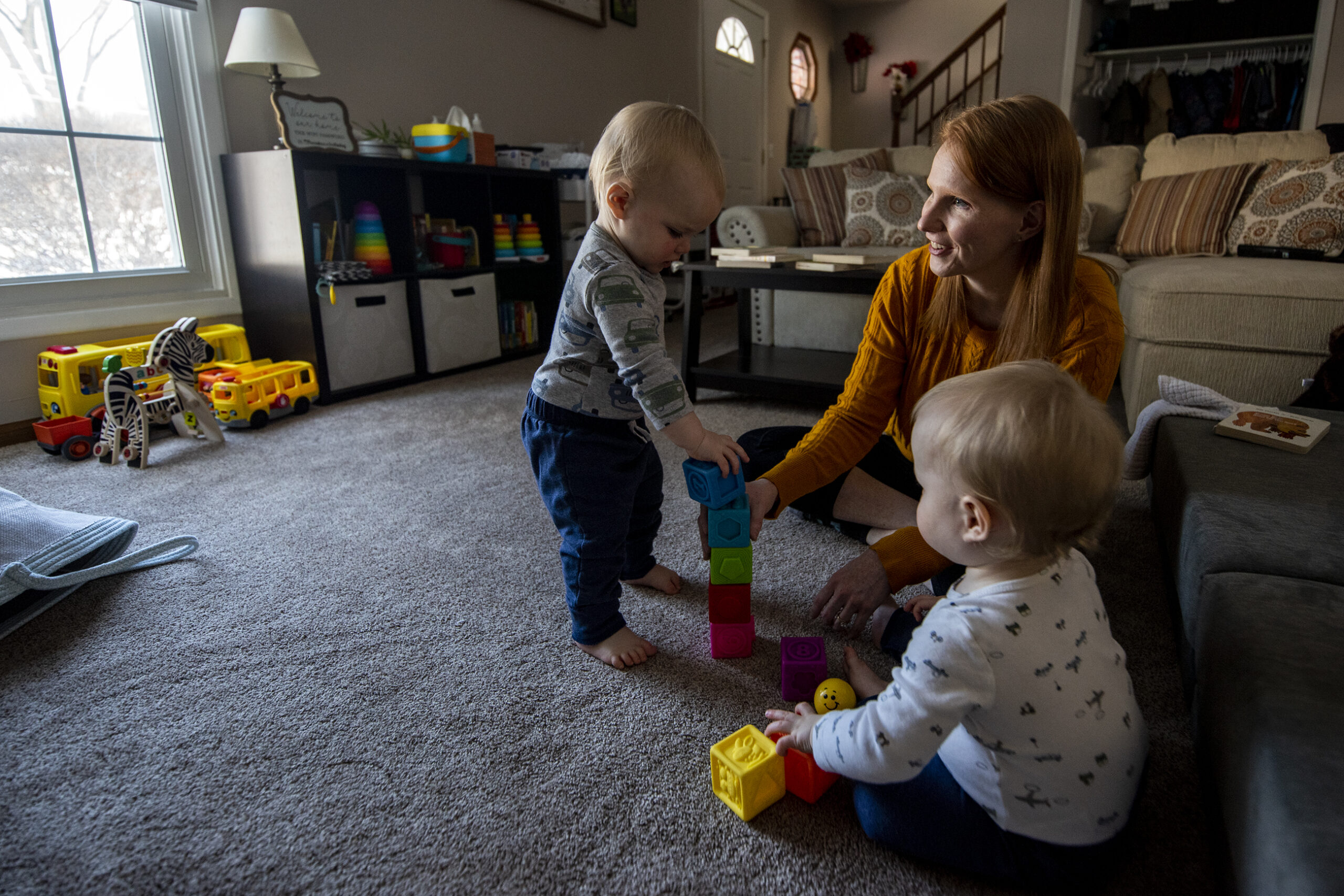 A woman sits on the ground with small twin boys as they play with colorful blocks.