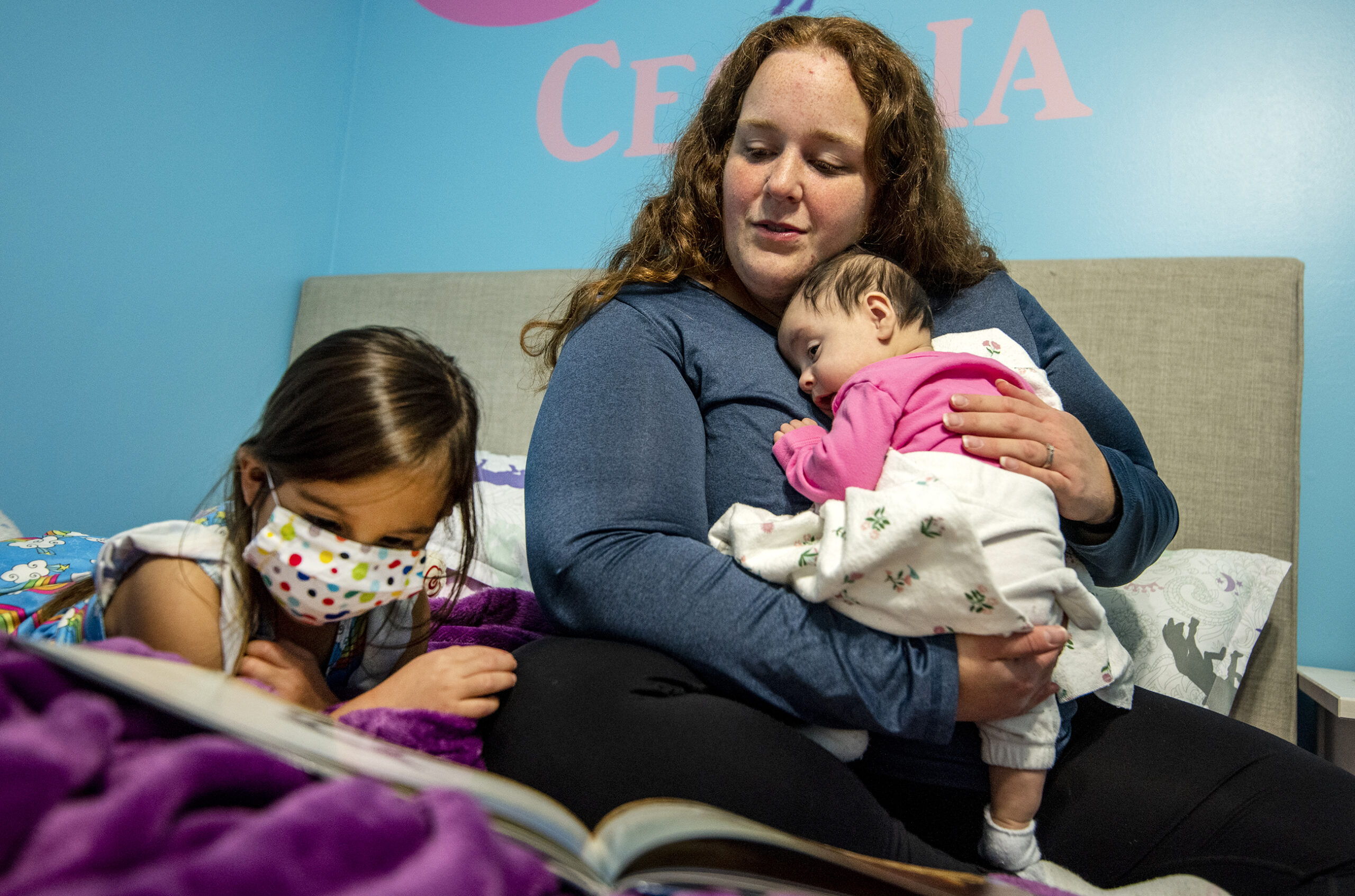 A woman holds a baby as a slightly older child reads from a book on her bed.