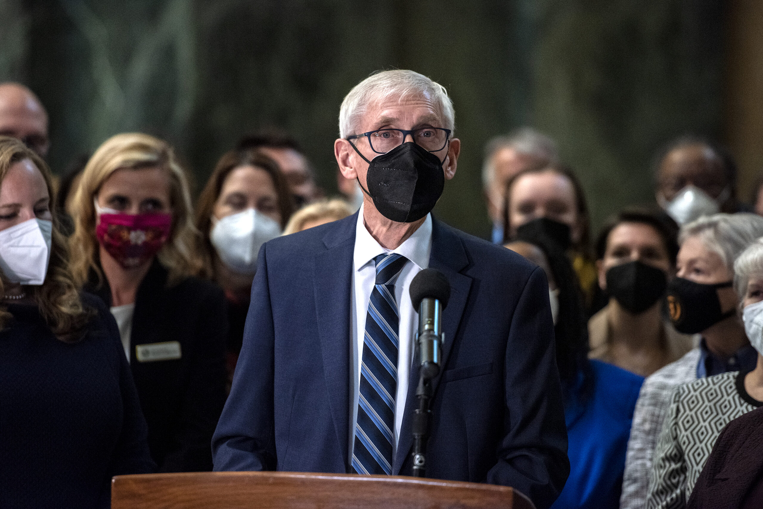 Gov. Tony Evers wears a black face mask as he speaks at a podium surrounded by lawmakers.
