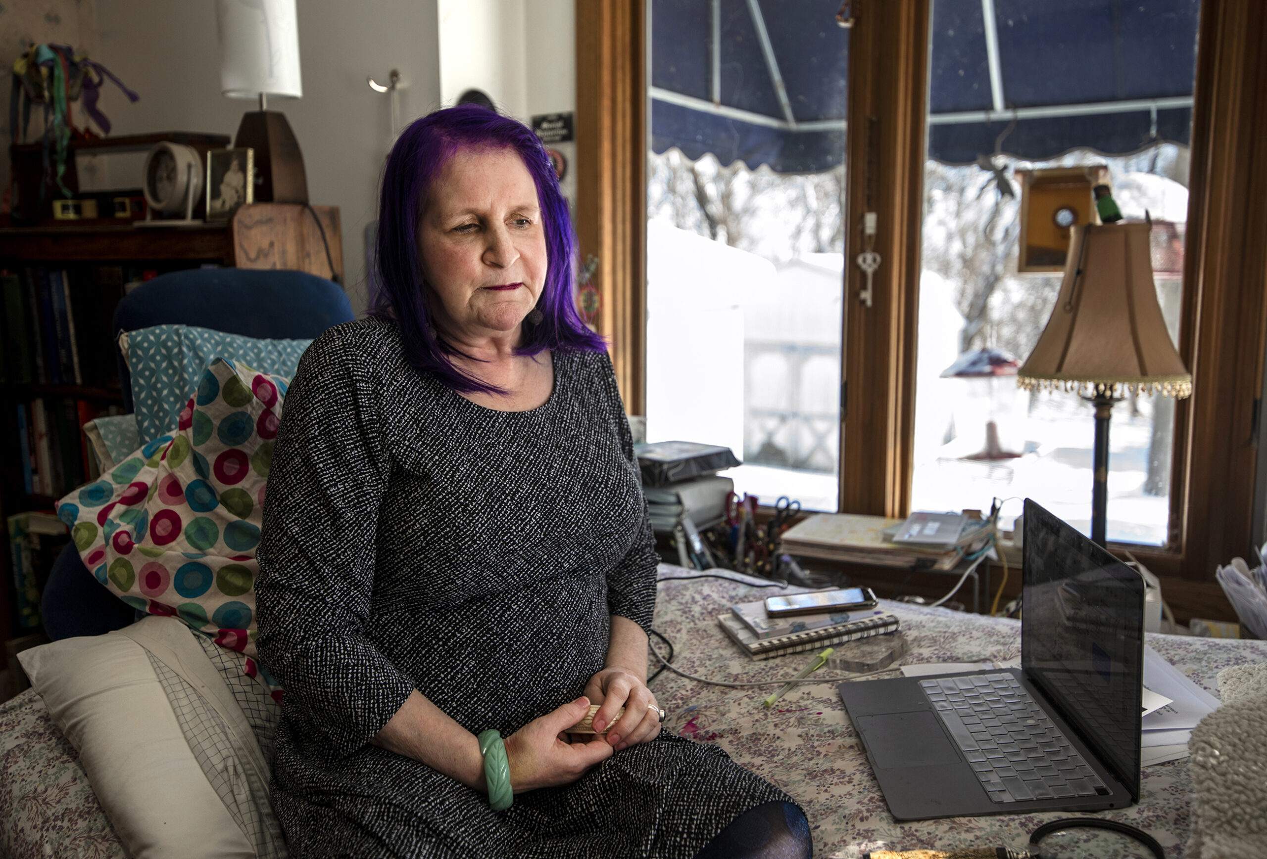 Sheryl Lamoureux looks down as she sits on a bed situated near windows.
