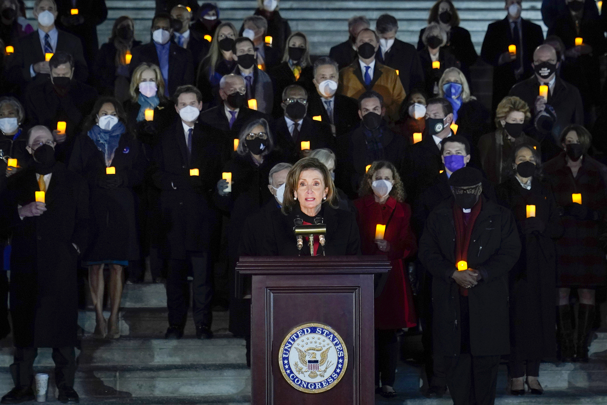 Nancy Pelosi speaks at a vigil on the anniversary of the Jan. 6 Capitol insurrection, with a crowd standing behind her and holding candles