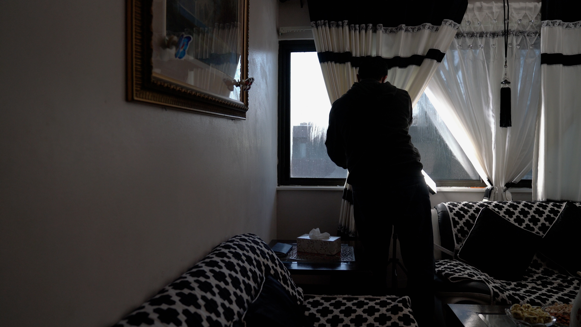 Khalid stands behind curtains framing a window and between couches inside an apartment.