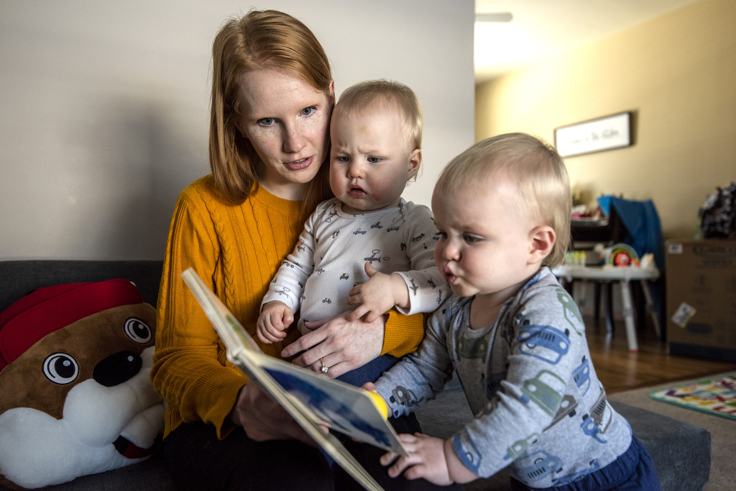A woman in a yellow sweater sits with twin boys on her lap as they read a book.
