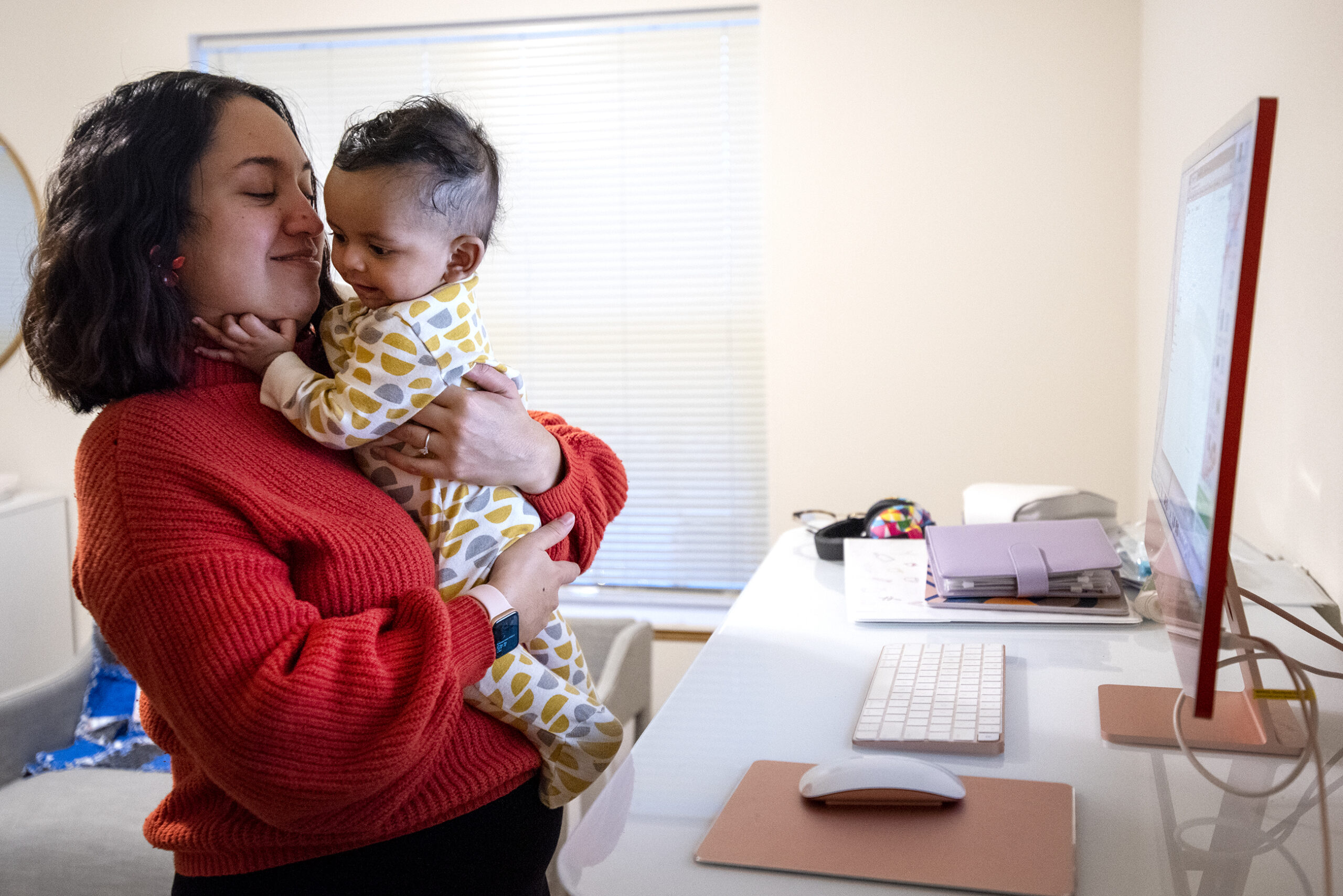 A woman holds a baby as she stands near a desk with a computer.