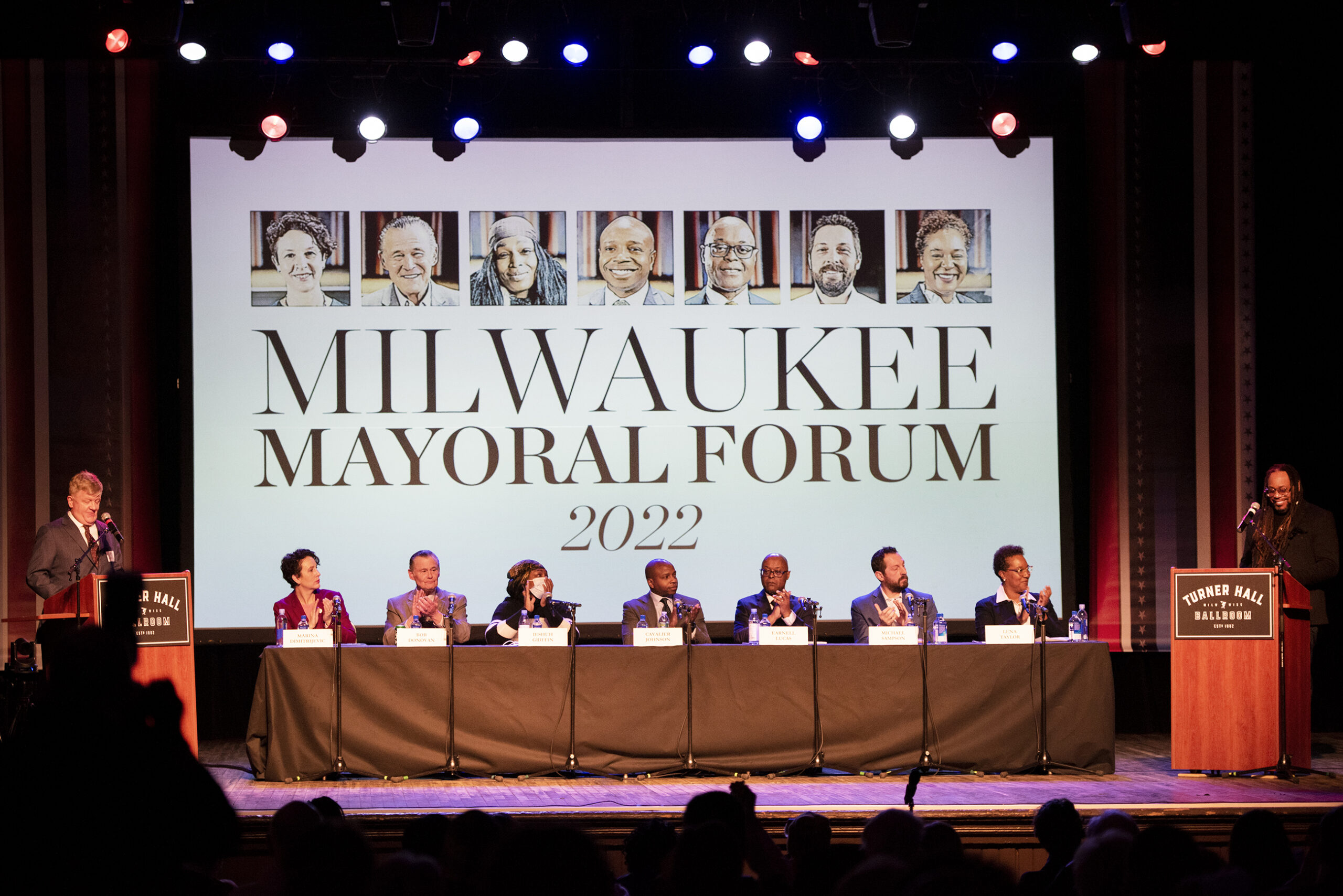 Seven candidates sit at a table on a theater stage in front of a screen that says "Milwaukee Mayoral Forum 2022."