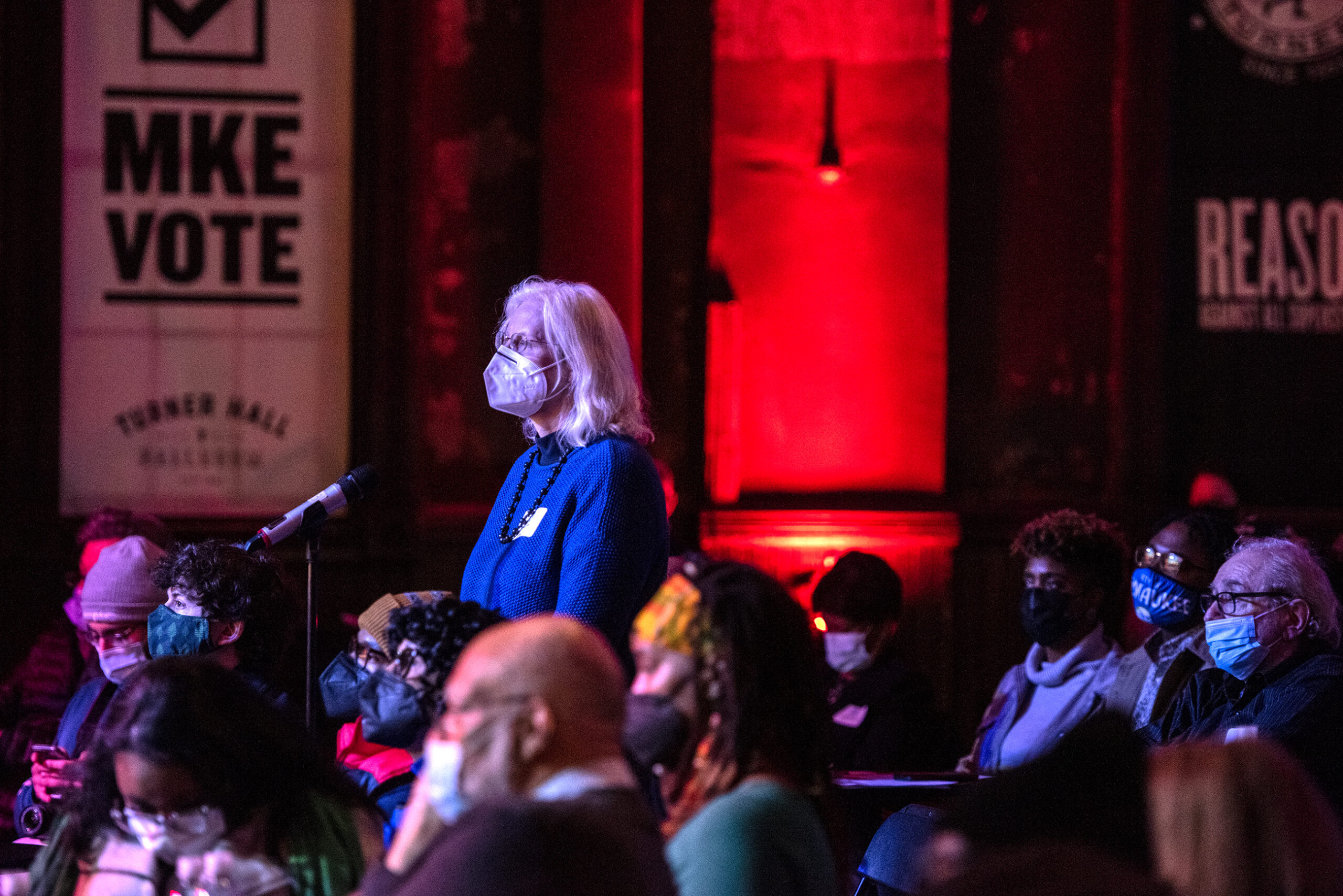 An attendee stands at a microphone to ask a question. She's surrounded by other seated members of the crowd.