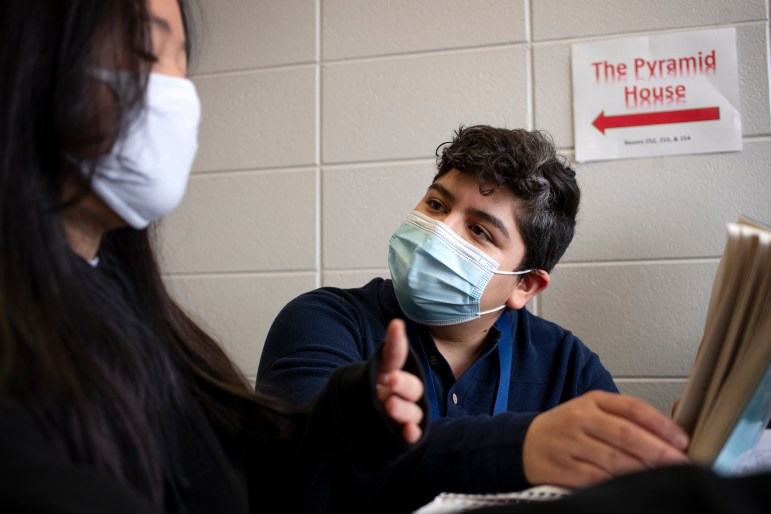 A boy wearing a mask reads a book next to a teacher