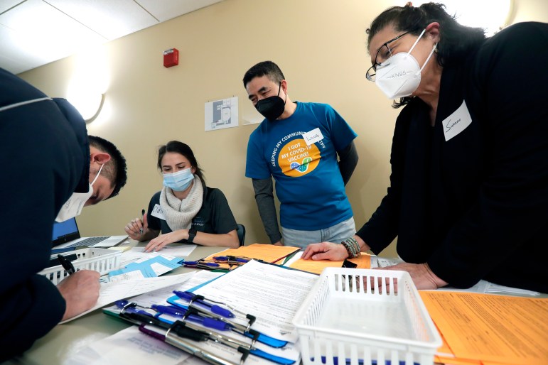 Three people stand at a table, registering another for a COVID-19 vaccine