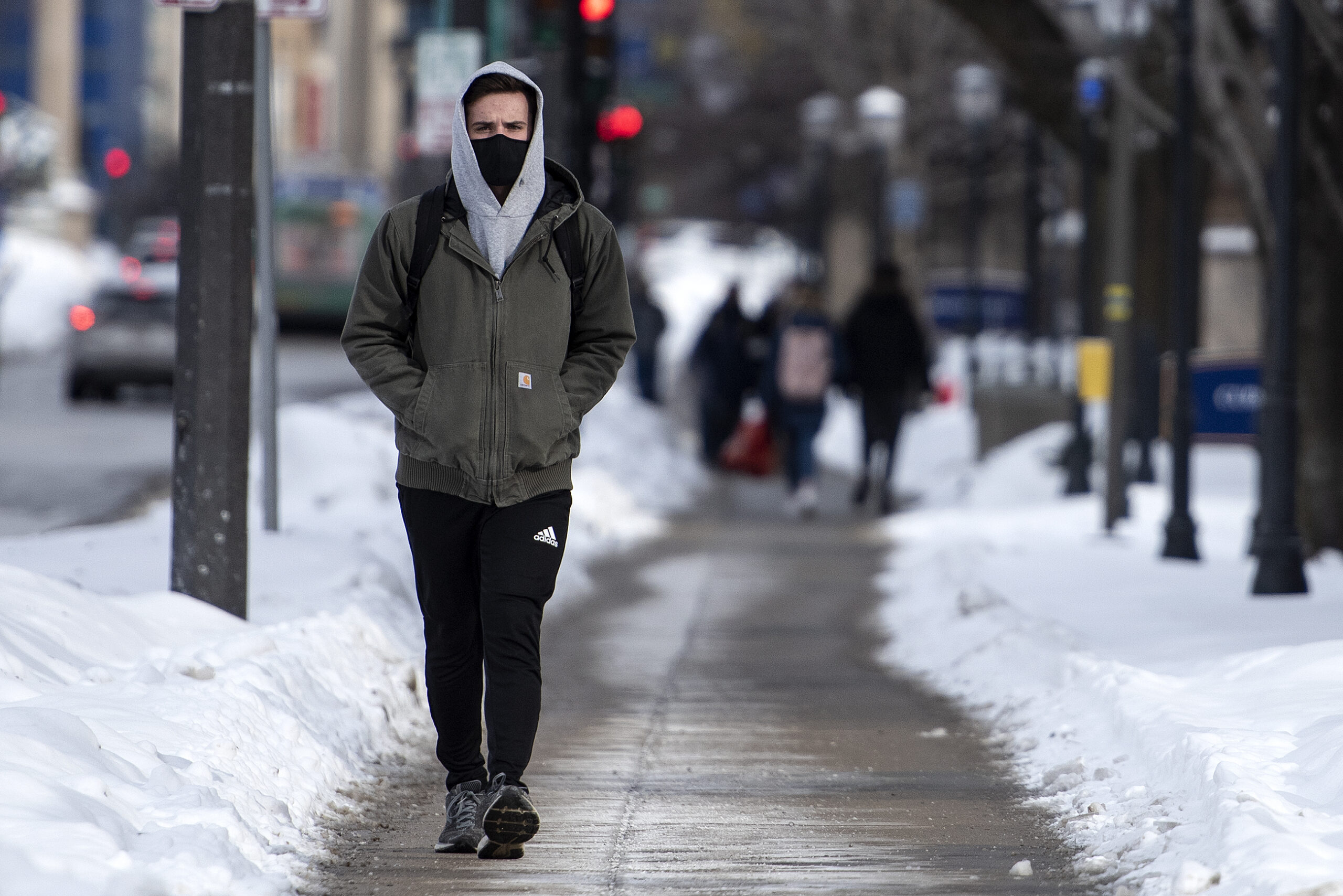 A man in winter clothing walks on a sidewalk flanked by snow.