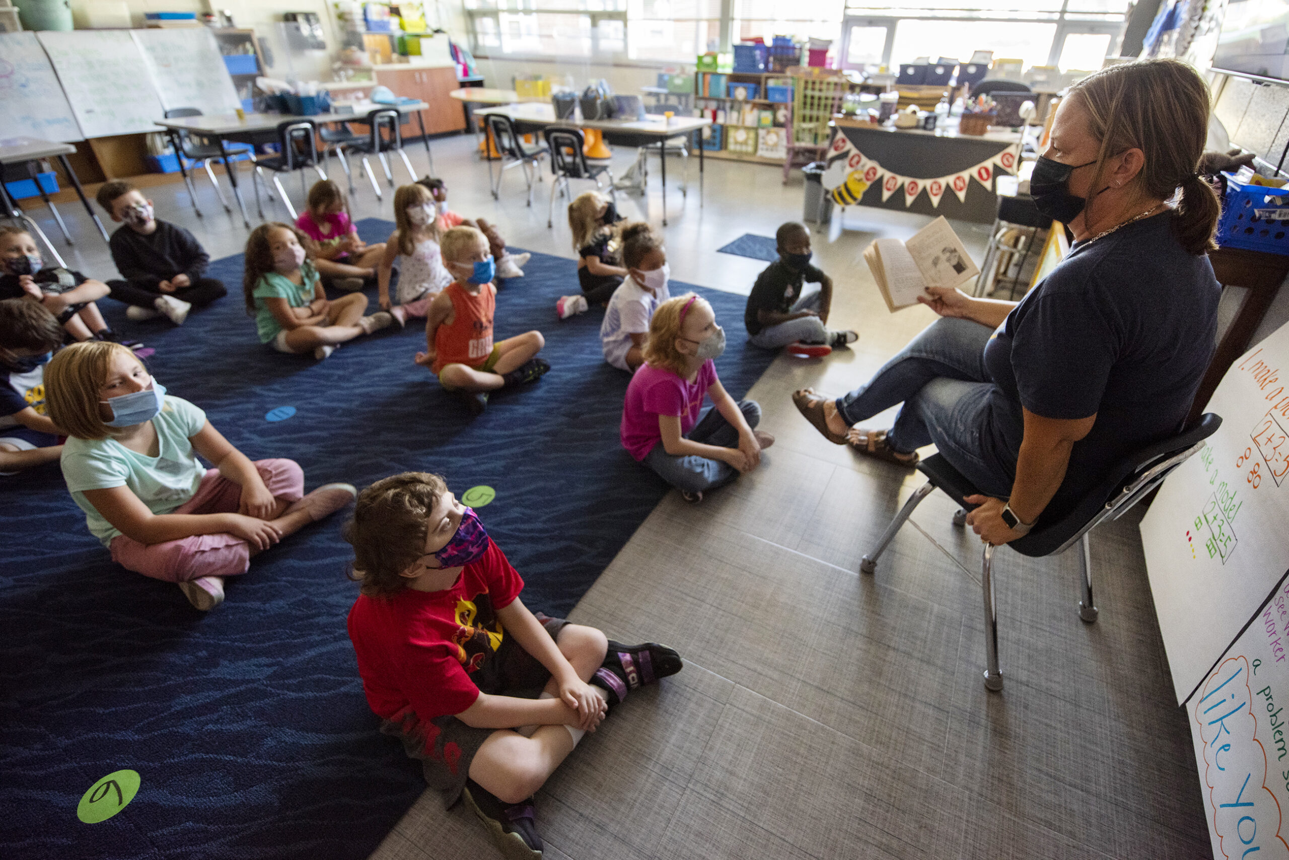 Children sit on a blue rug as a teacher reads a story.