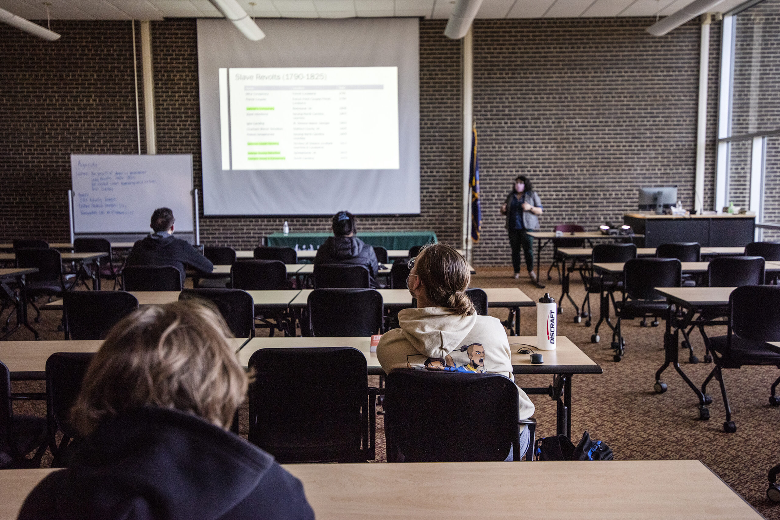 Several students sit at long desks in a lecture hall. A teacher at the front speaks as material appears on a screen.