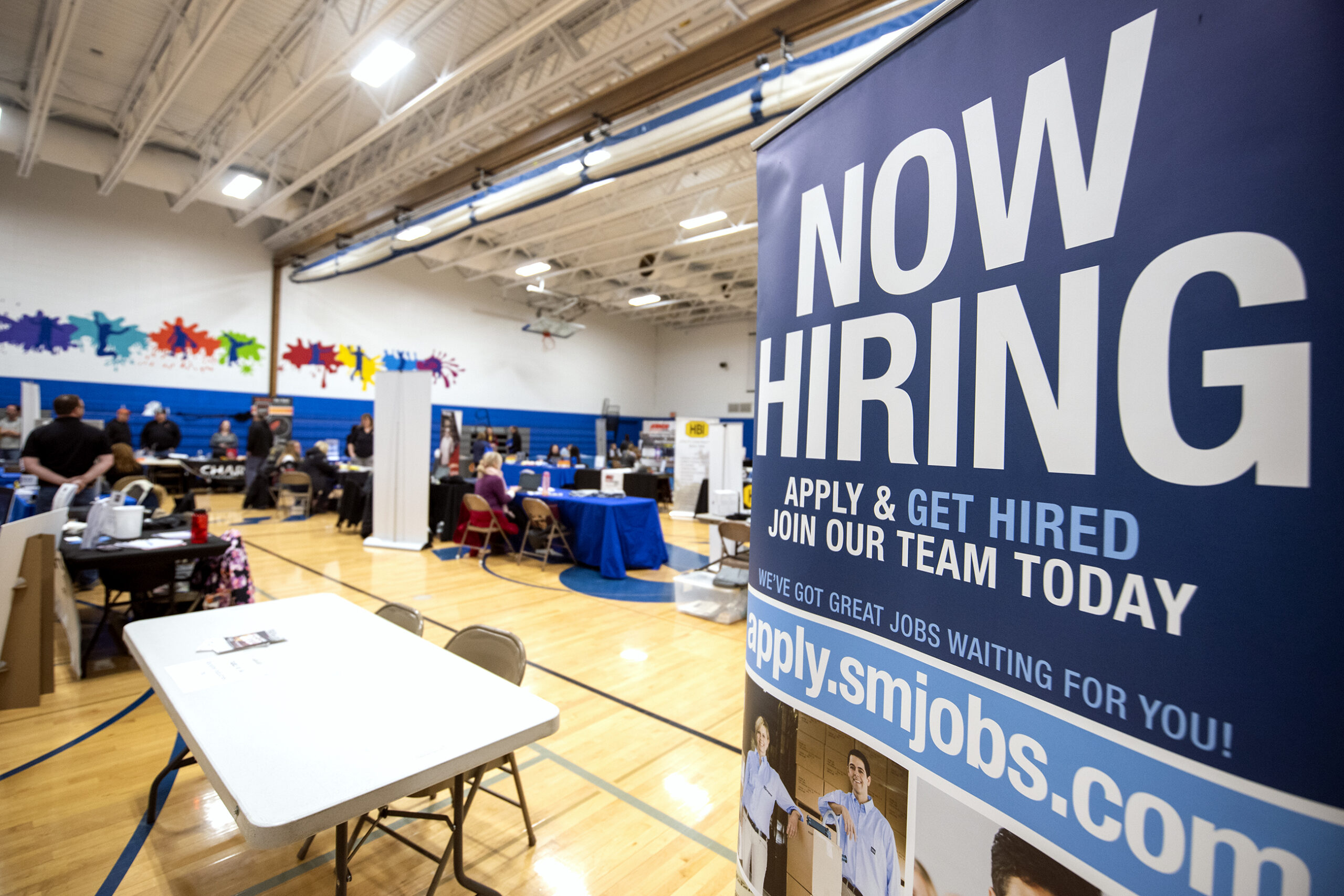 A blue banner at a career fair booth says "Now Hiring."